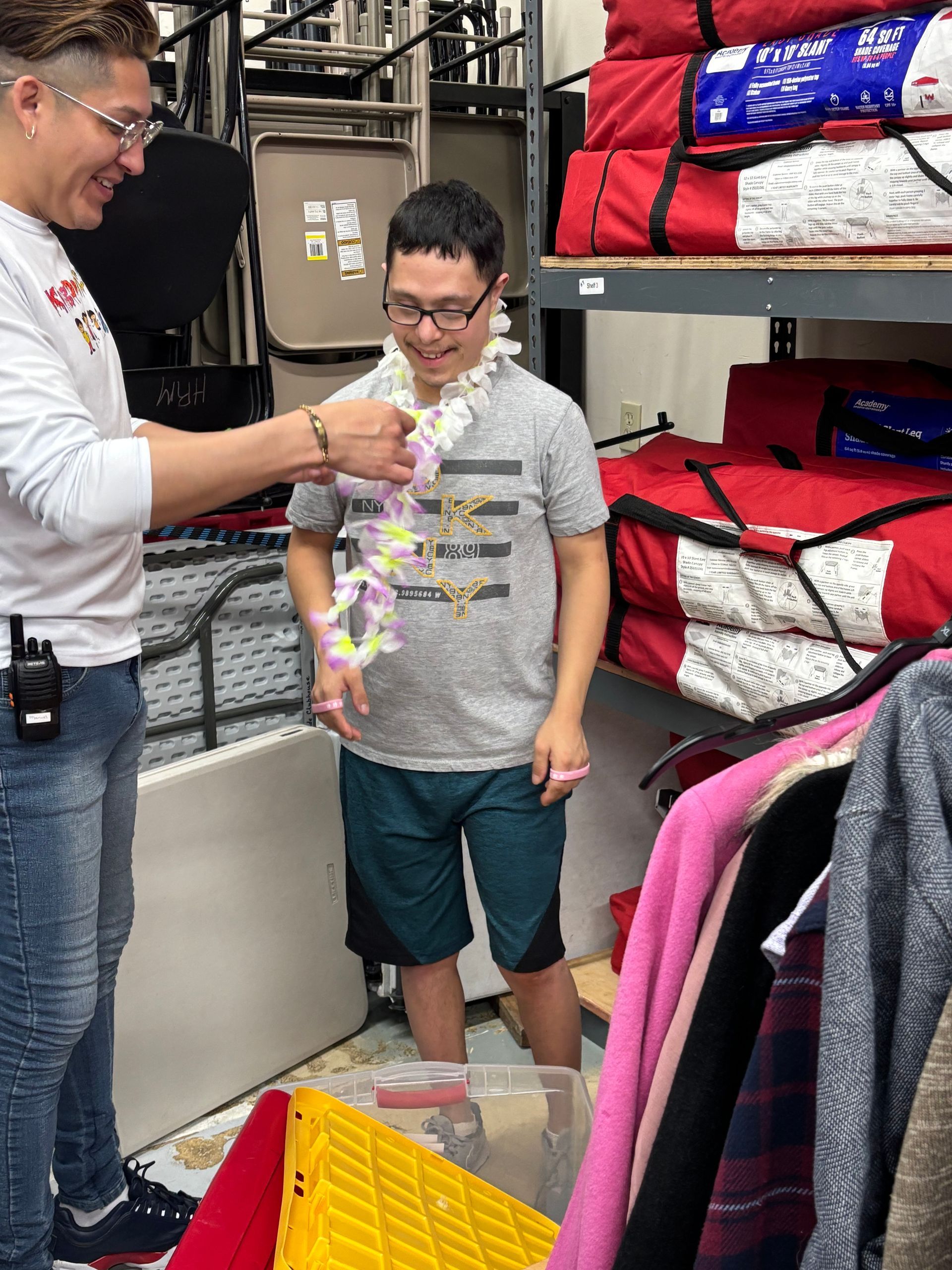 Man puts lei around another man's neck indoors. Shelves filled with items, including red bags.