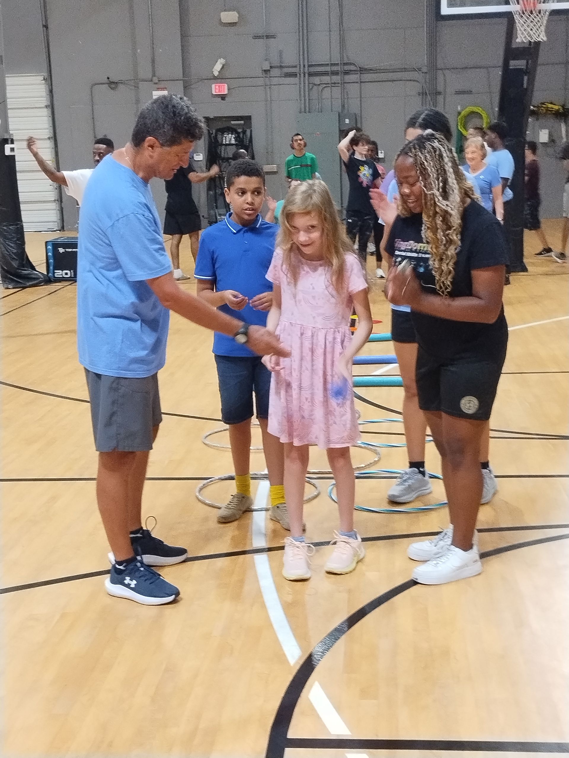 A man in blue shirt guides a young girl in a pink dress through agility hoops at a gym. Others watch.