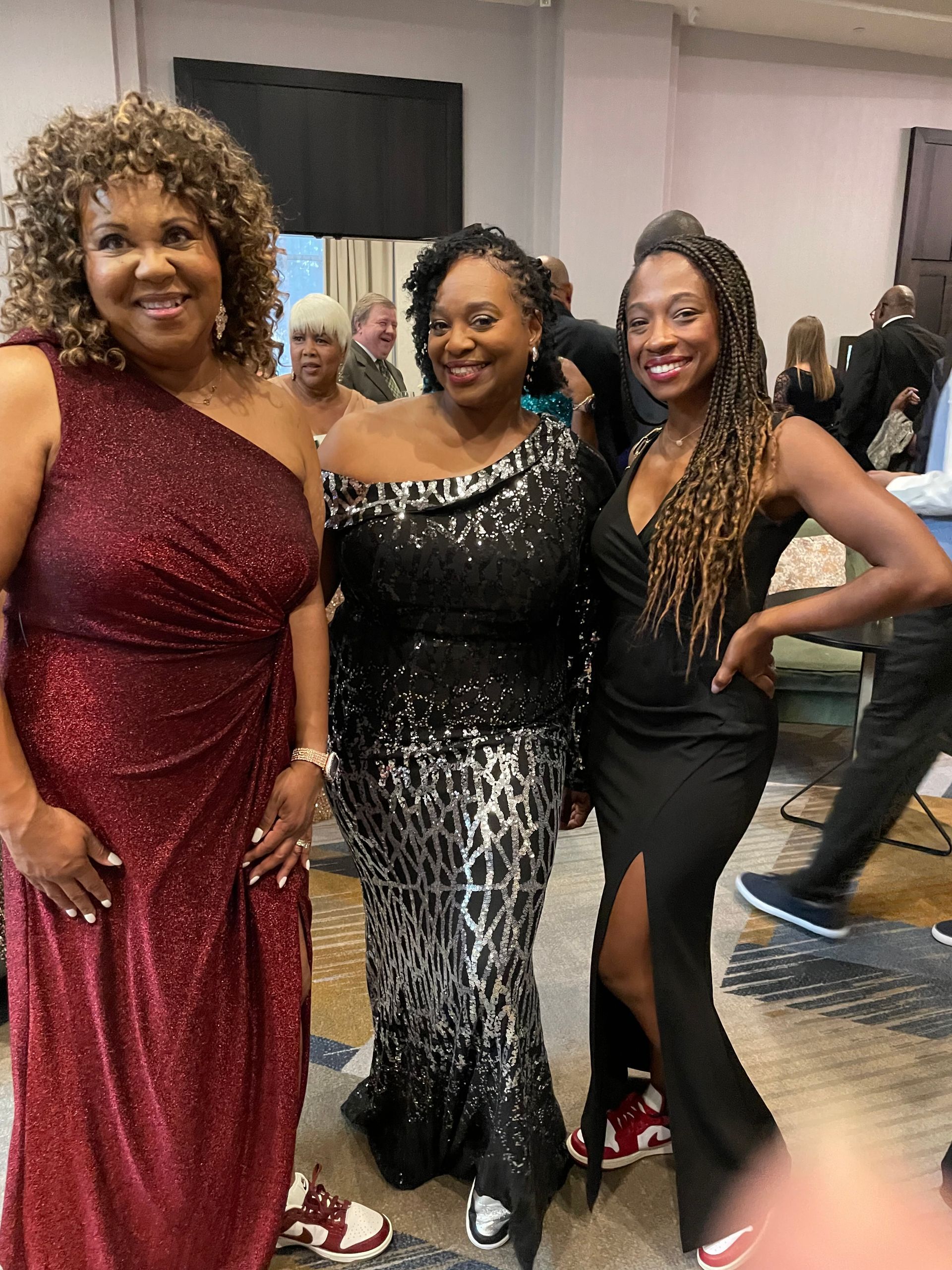 Three women in formal dresses pose at an event—red sequined, black and silver print, and black with a slit.