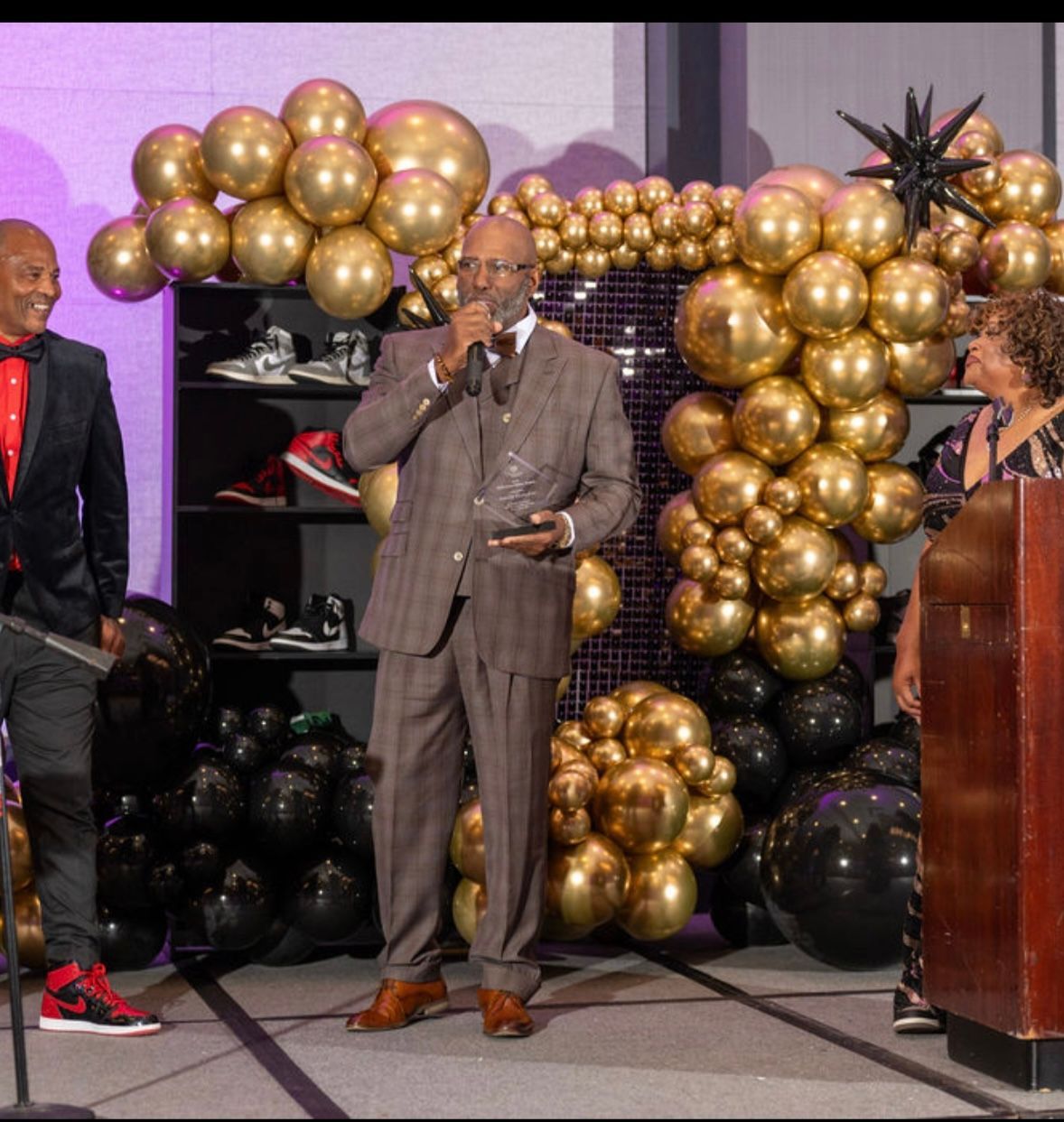 Man in suit accepting award, standing at podium with gold and black balloon decorations. Two people stand nearby.