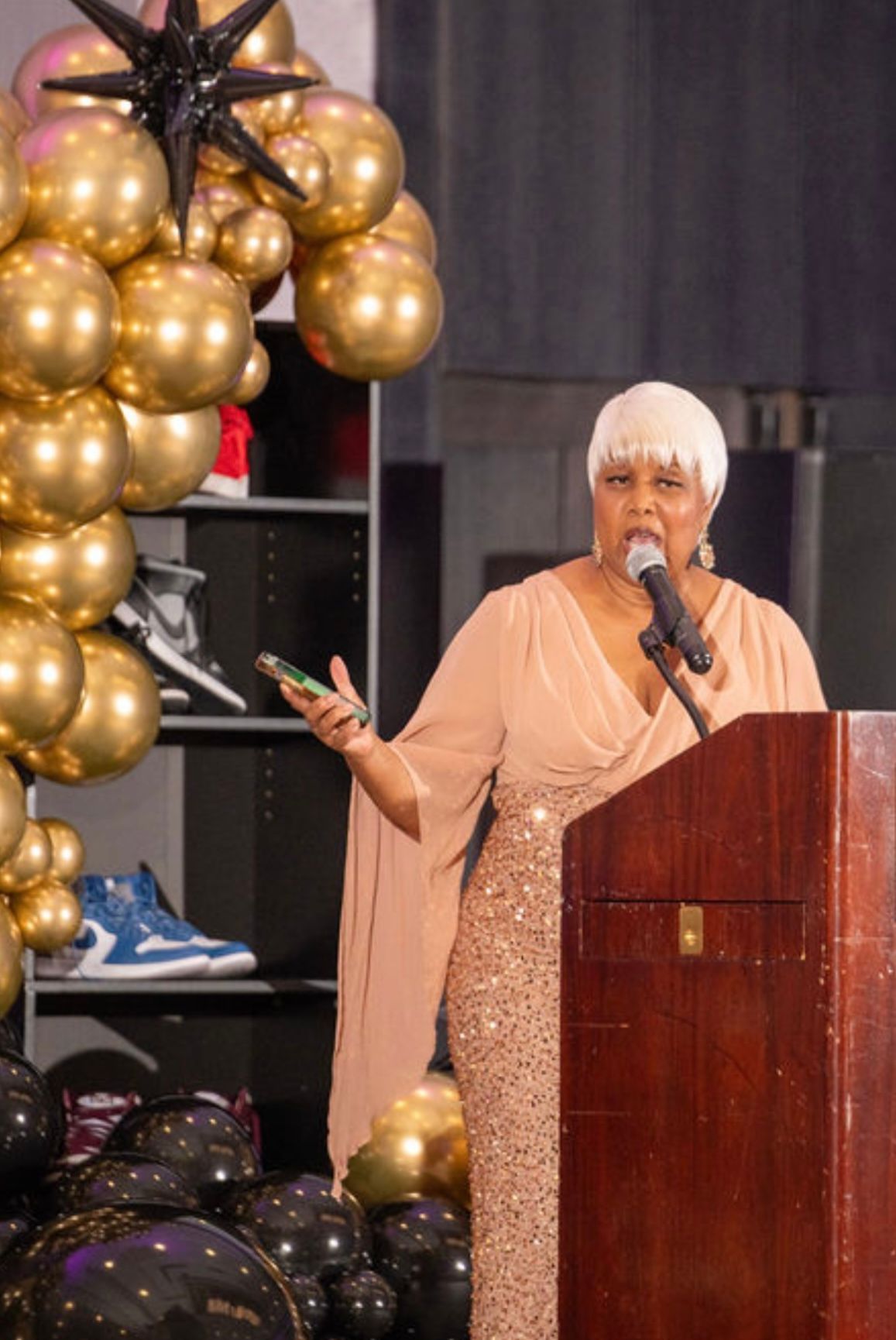 Woman in a shimmering gown speaks at a podium, with golden balloons and a sneaker display in the background.