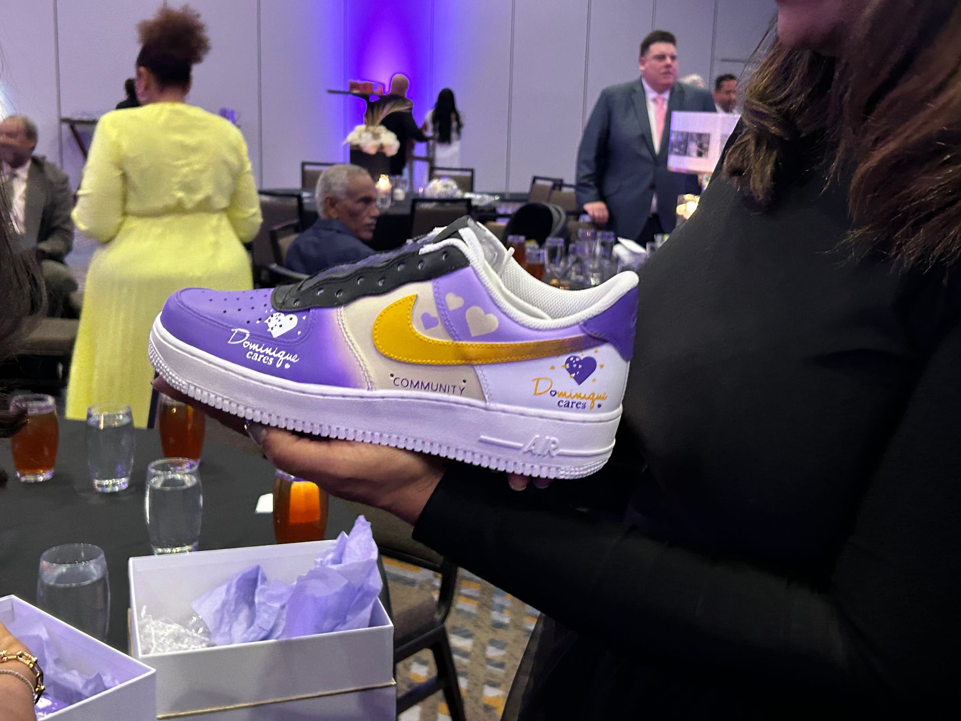 Woman holding a custom purple and gold Nike Air Force 1 sneaker at an event.