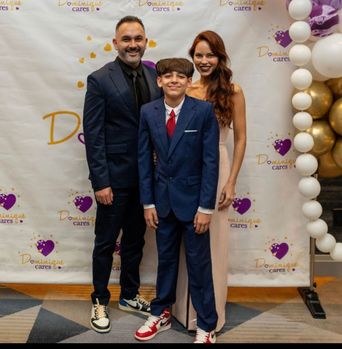 Family poses at an event. Man in suit, boy in suit, woman in gown smile near a backdrop with balloons.