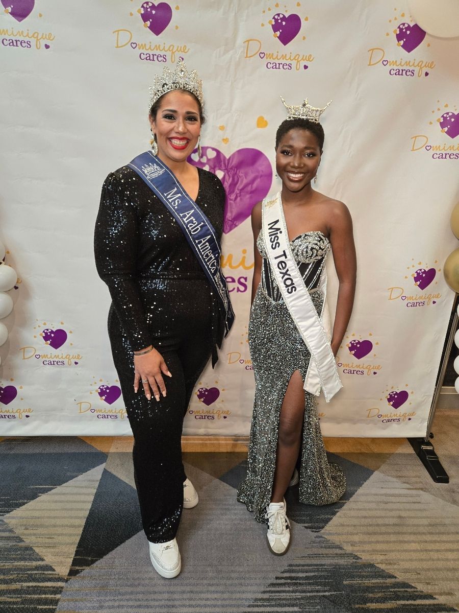 Two women in crowns and sashes at an event, standing near a purple heart banner.