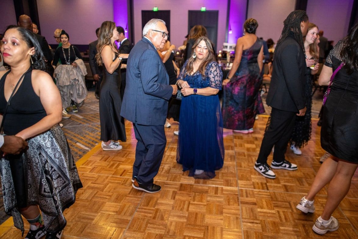People in formal attire at a gala, dancing on a wooden floor, illuminated by purple lights.