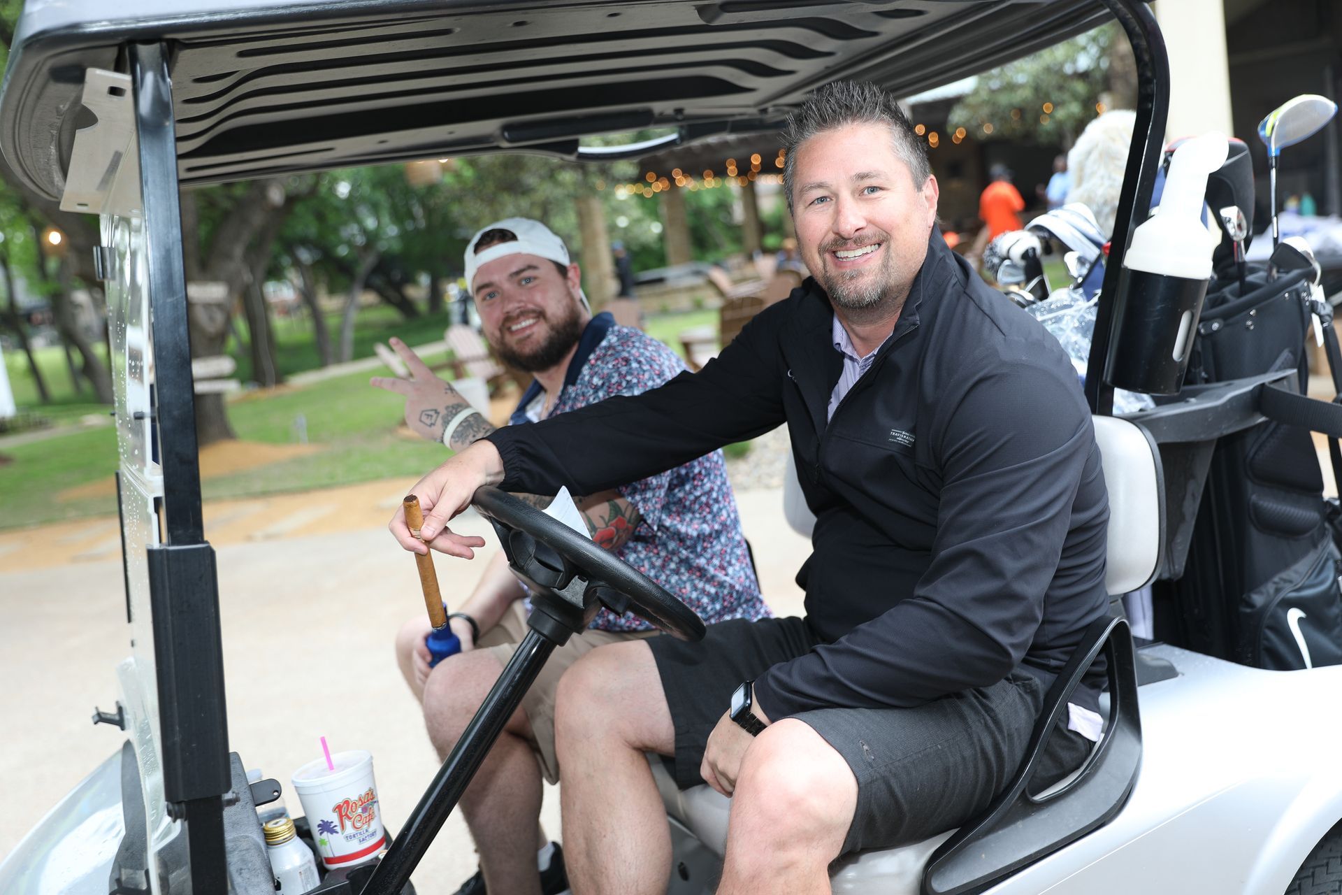 Two men in a golf cart. One drives, the other waves. Outdoors, trees in the background.