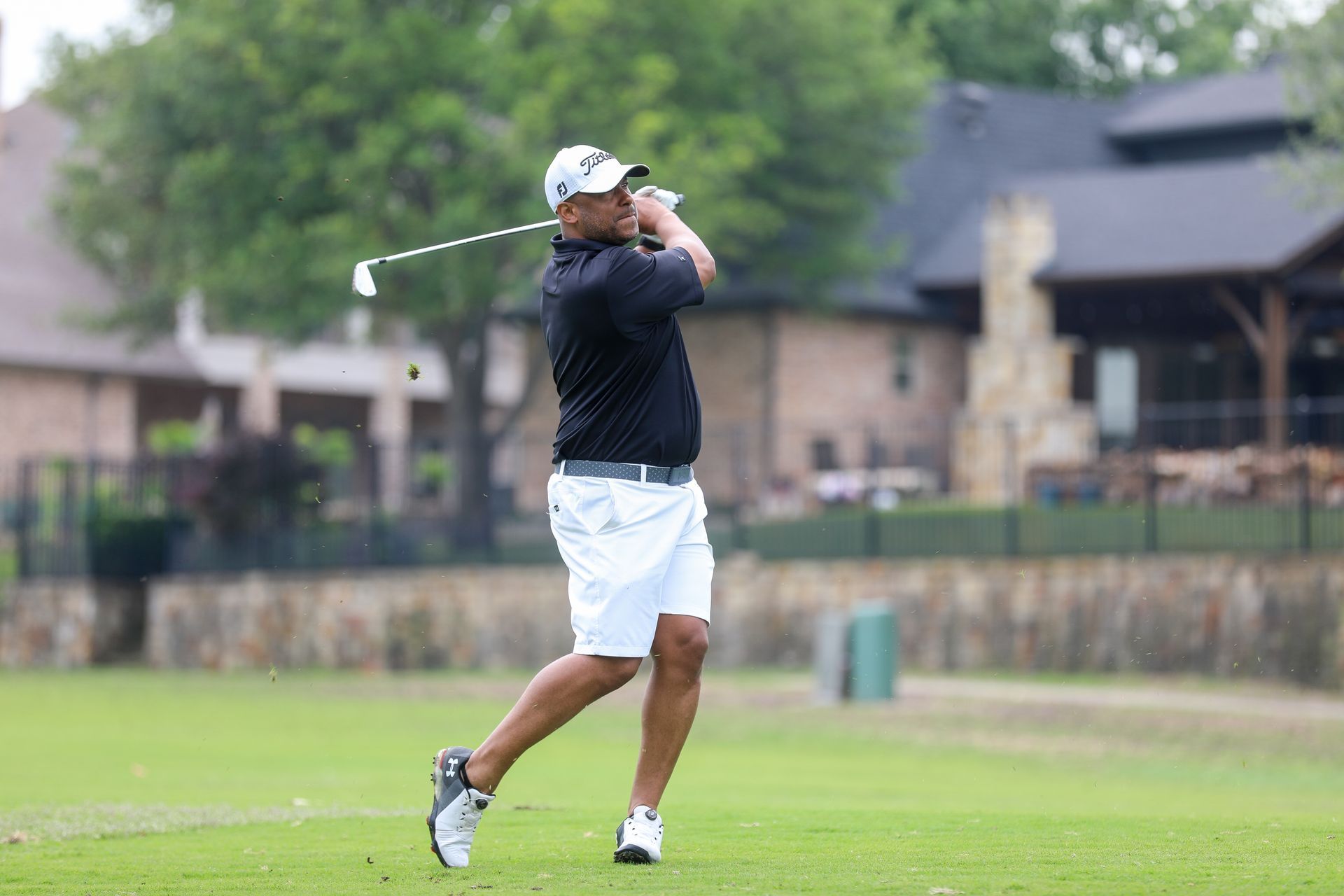 Golfer in white shorts and black shirt swings club on green course near a building.