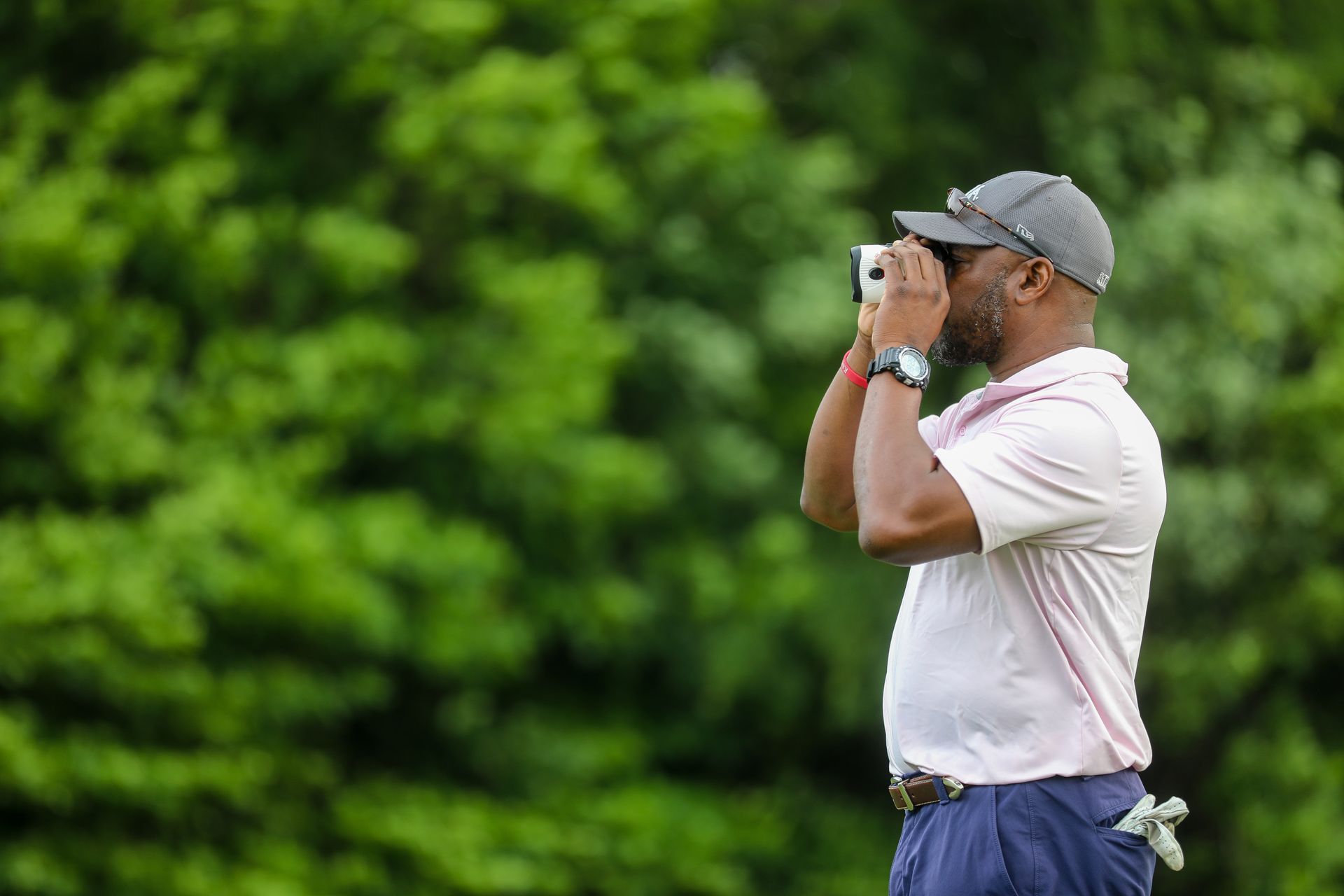 Man in golf attire looking through a rangefinder on a golf course, with green foliage in the background.