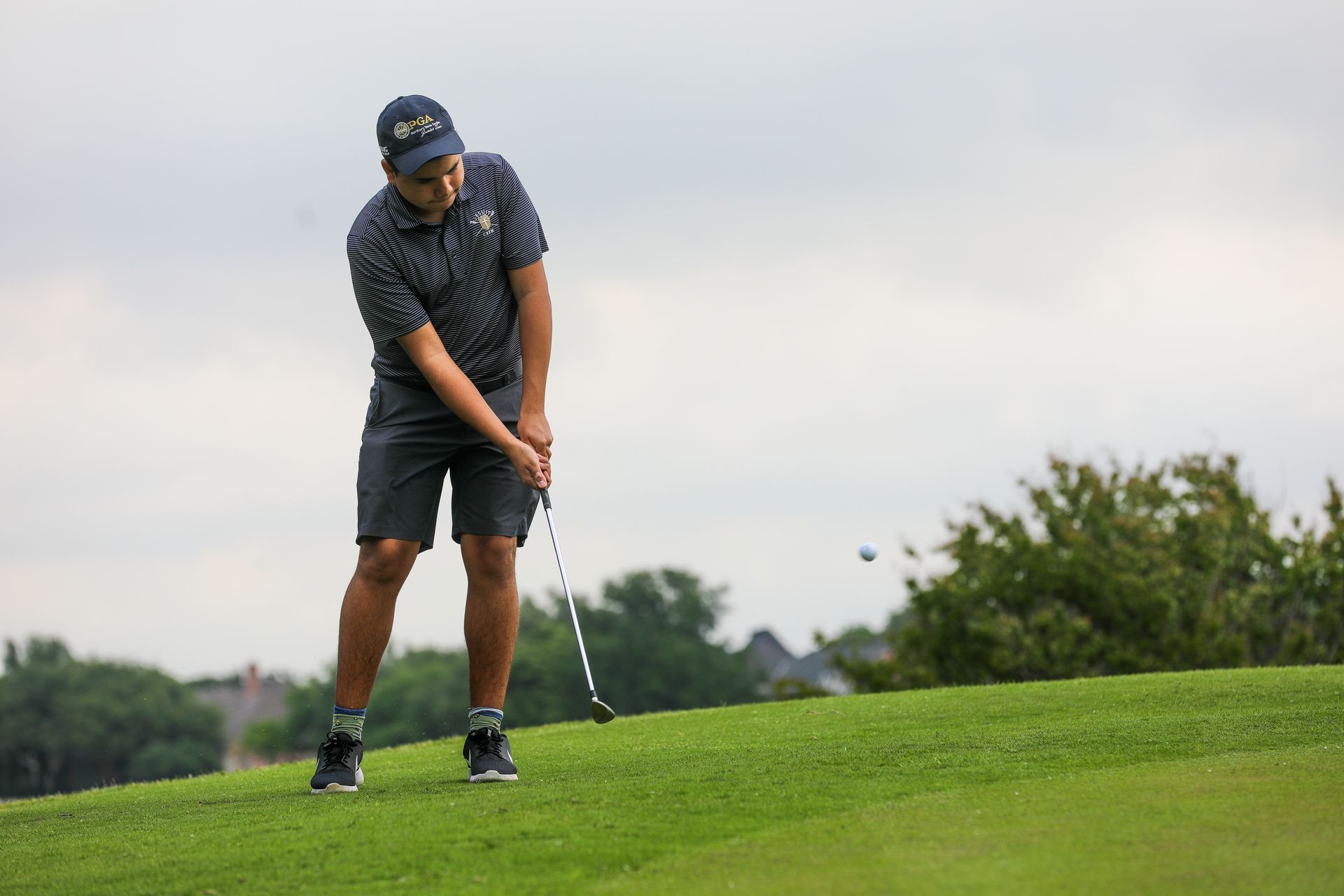 Golfer in a navy patterned shirt and shorts swings club on a green course under an overcast sky.