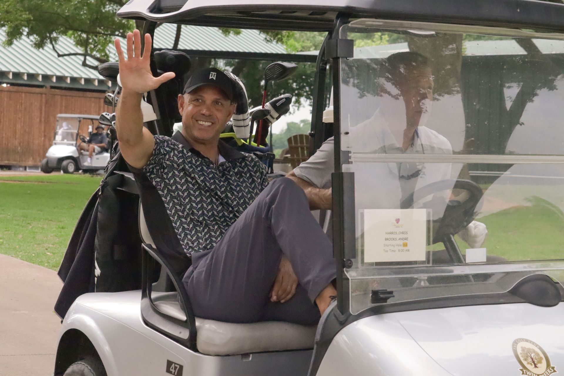 Man waving from a golf cart on a course. Wearing a patterned shirt and cap.