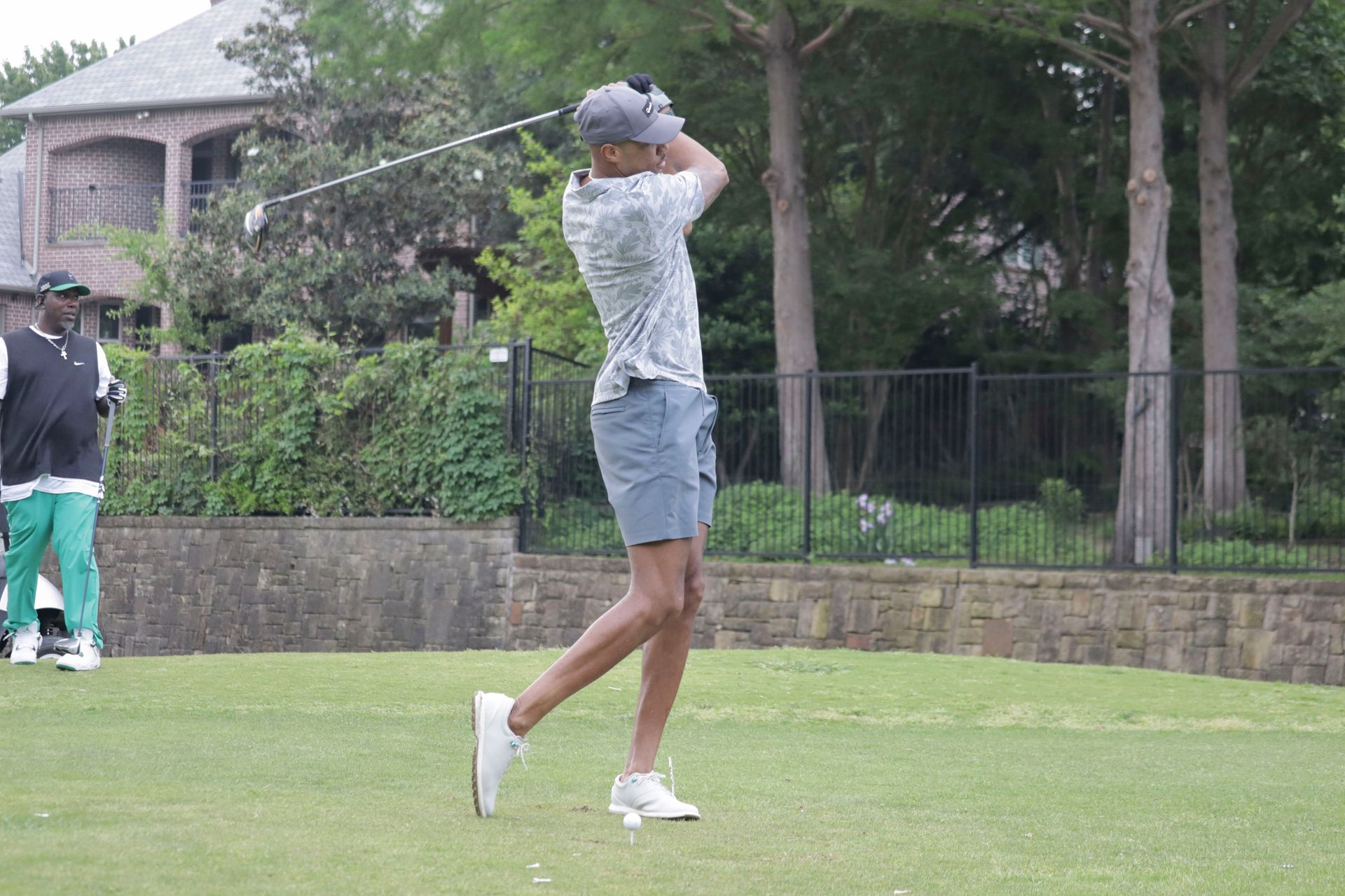 Man in golf swing on green, gray shorts and shirt, white shoes, brick wall, trees, and house in background.