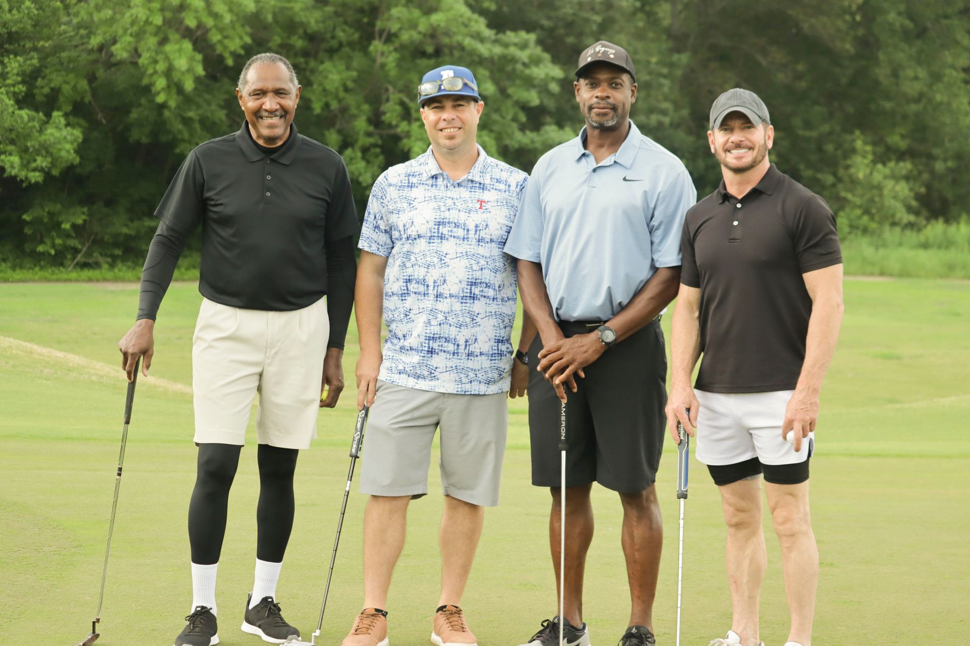 Four men on a golf course: two black and two white. Posing with clubs, green grass, sunny day.