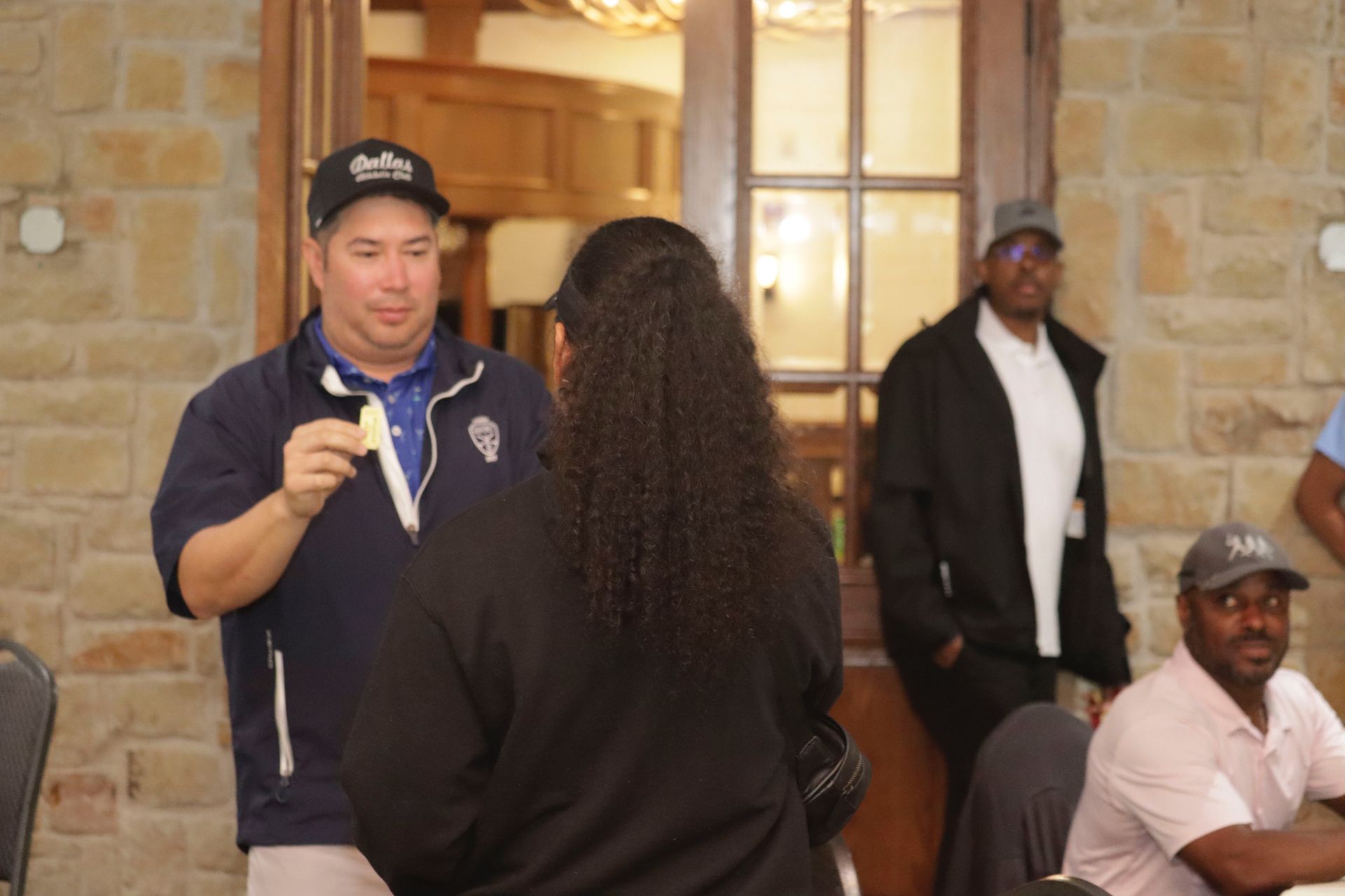 Man in golf attire talking to woman, others in a room, stone wall background.