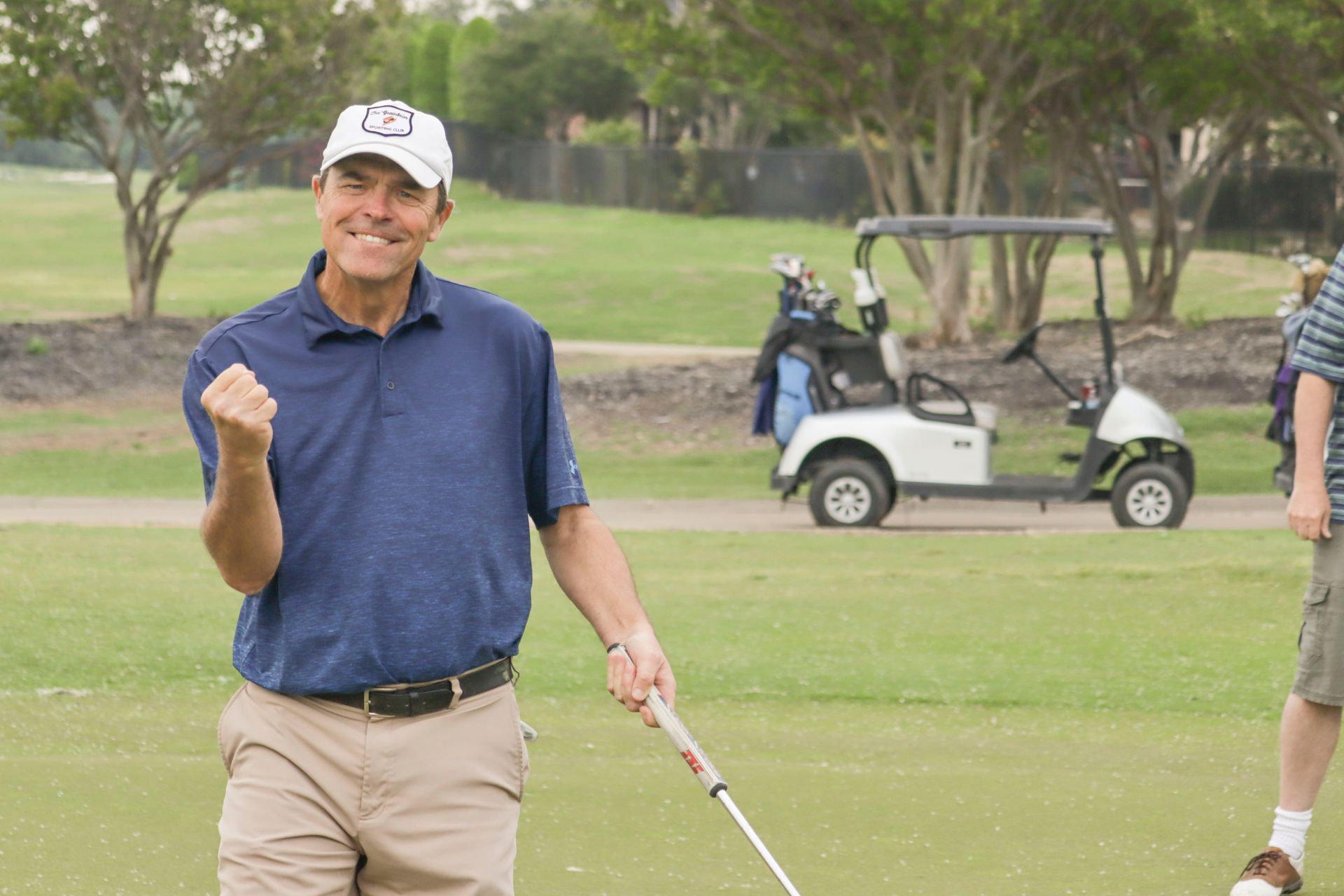 Man on a golf course celebrates with a raised fist, holding a club. He wears a white hat, blue shirt, and beige shorts.