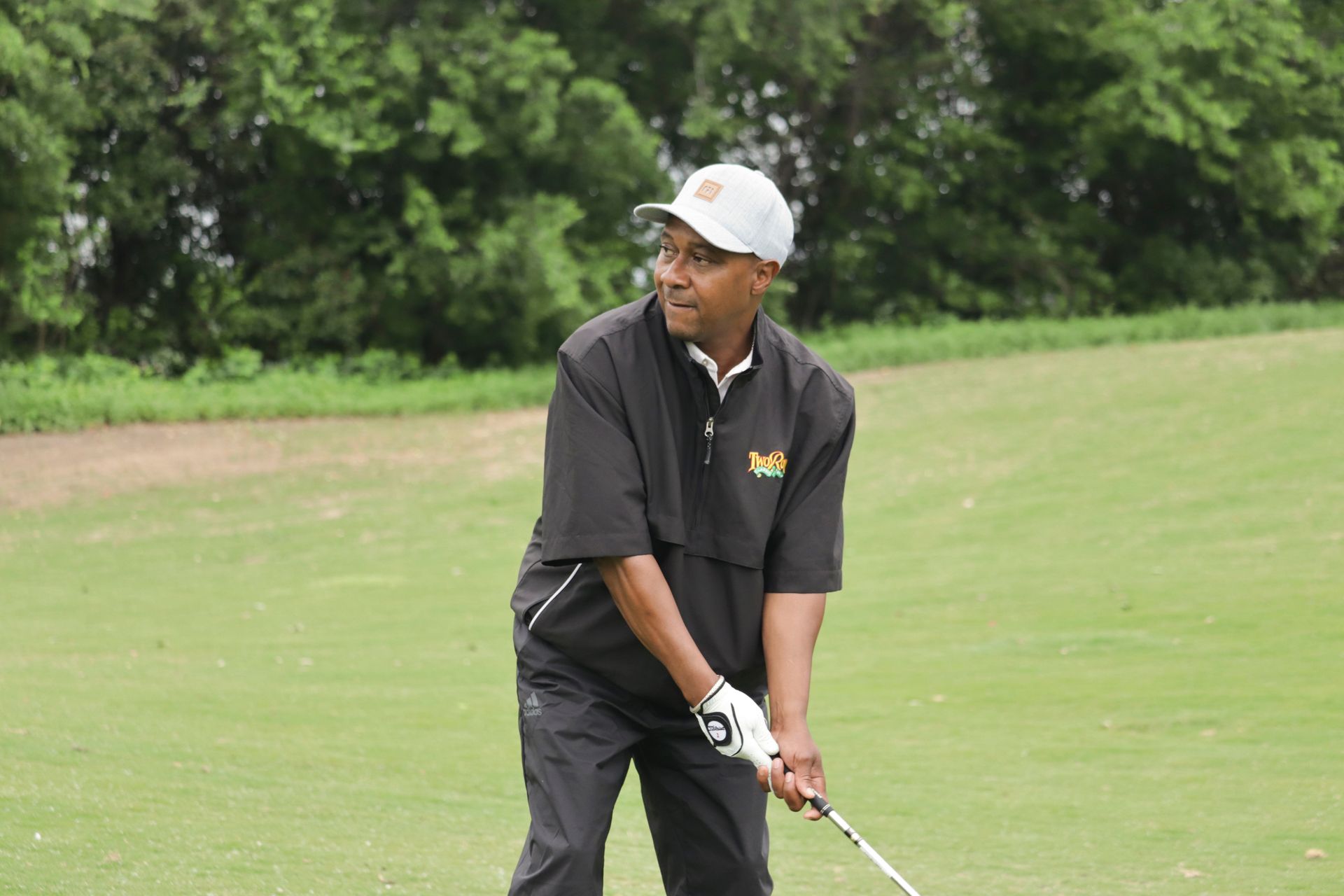 Man in black golf attire, wearing a white cap, swings a golf club on a green course.