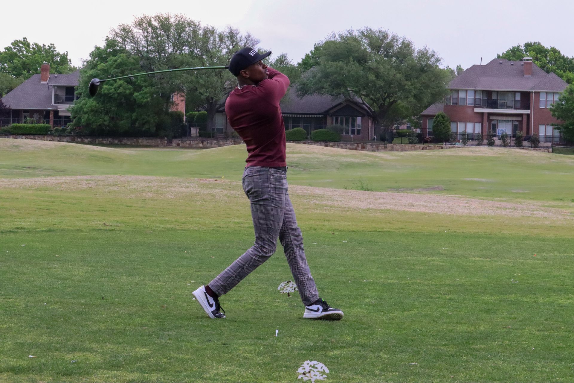Golfer swinging a club on a golf course; wearing maroon top, patterned pants, and white/black shoes.