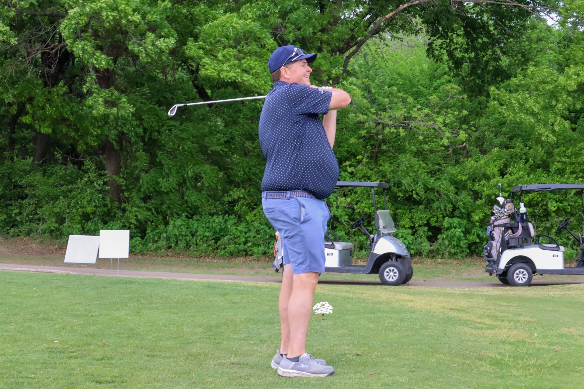 Man in blue shirt swings a golf club on a green, near golf carts and trees.