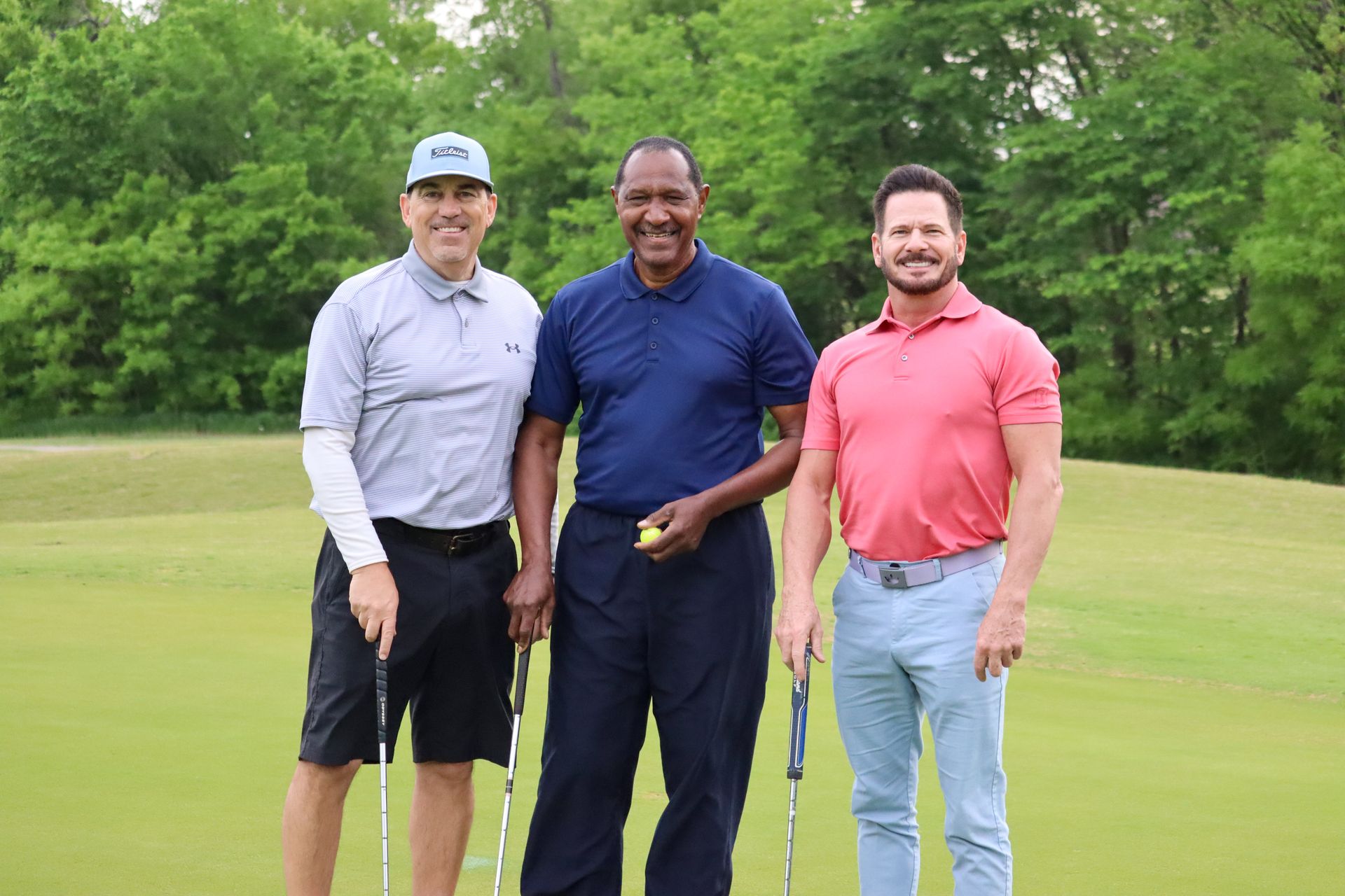 Three men smiling on a golf course; one holds a golf ball, others hold clubs. Green grass, trees in background.