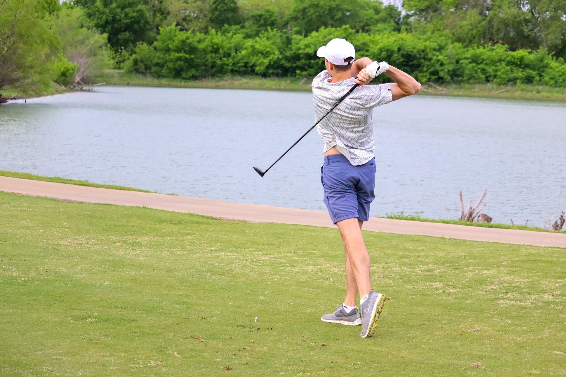Golfer in white hat mid-swing, facing away, blue shorts, green grass, lake background.