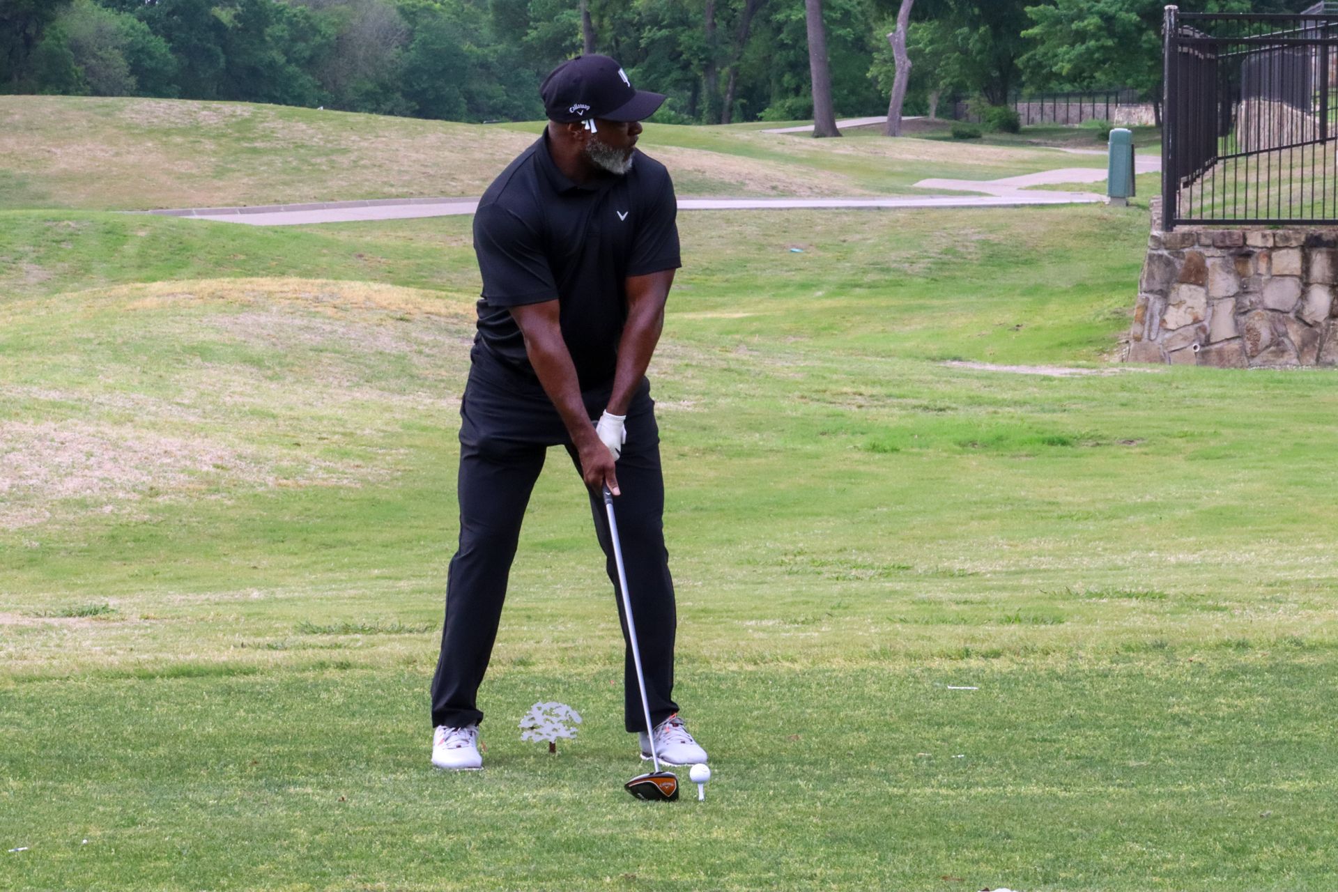 Man in black golf attire swings club on a grassy course, tee and ball in foreground.