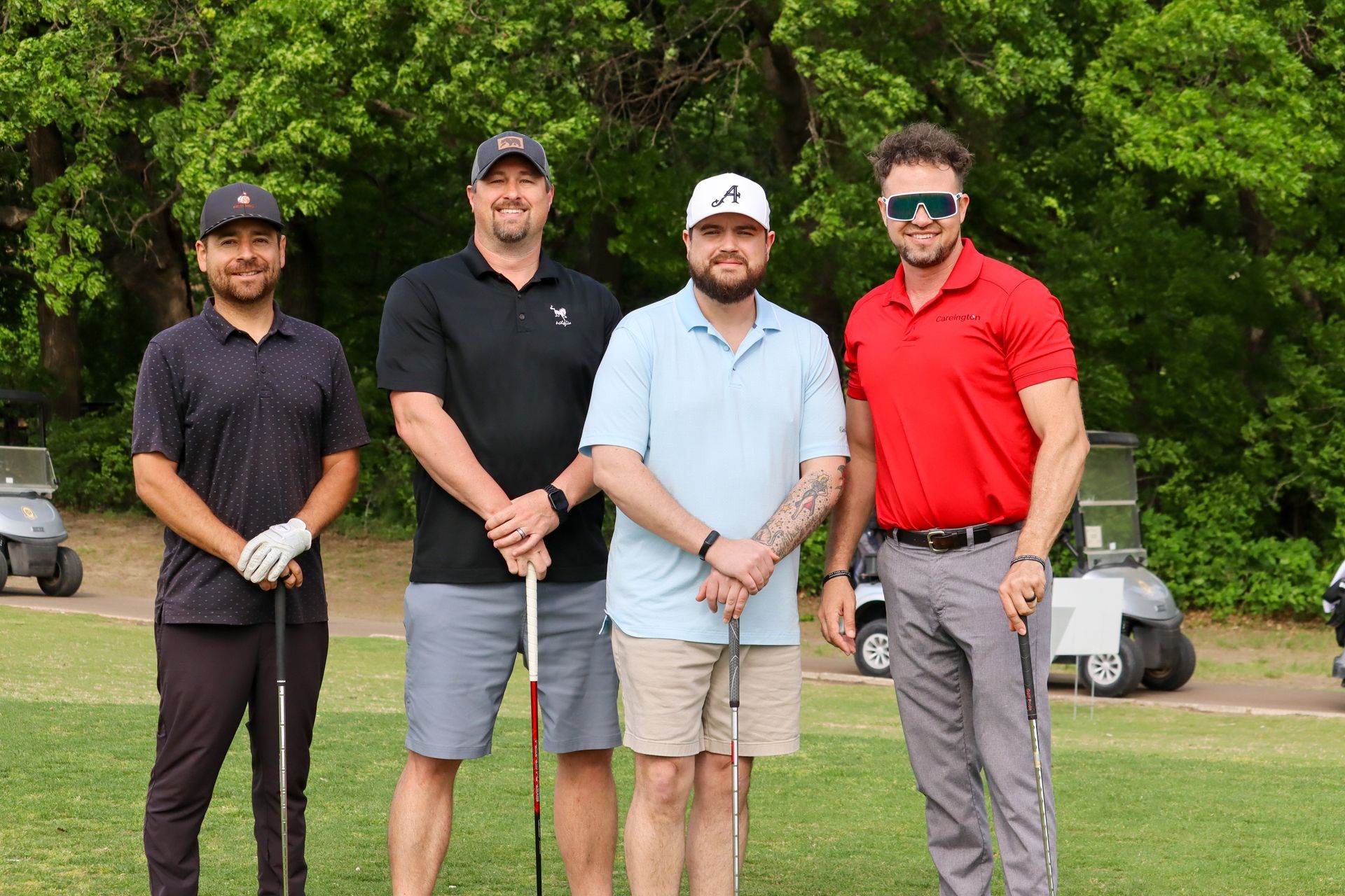 Four men standing on a golf course holding clubs, smiling. Two golf carts are in the background.