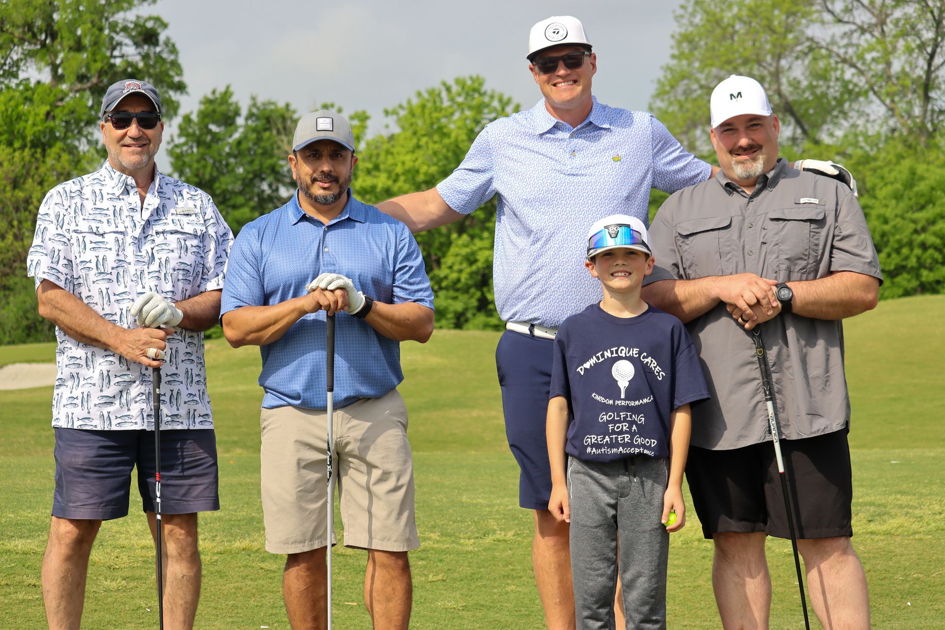 Five people on a golf course, posing with clubs. Men and a boy smiling, green grass and trees in the background.
