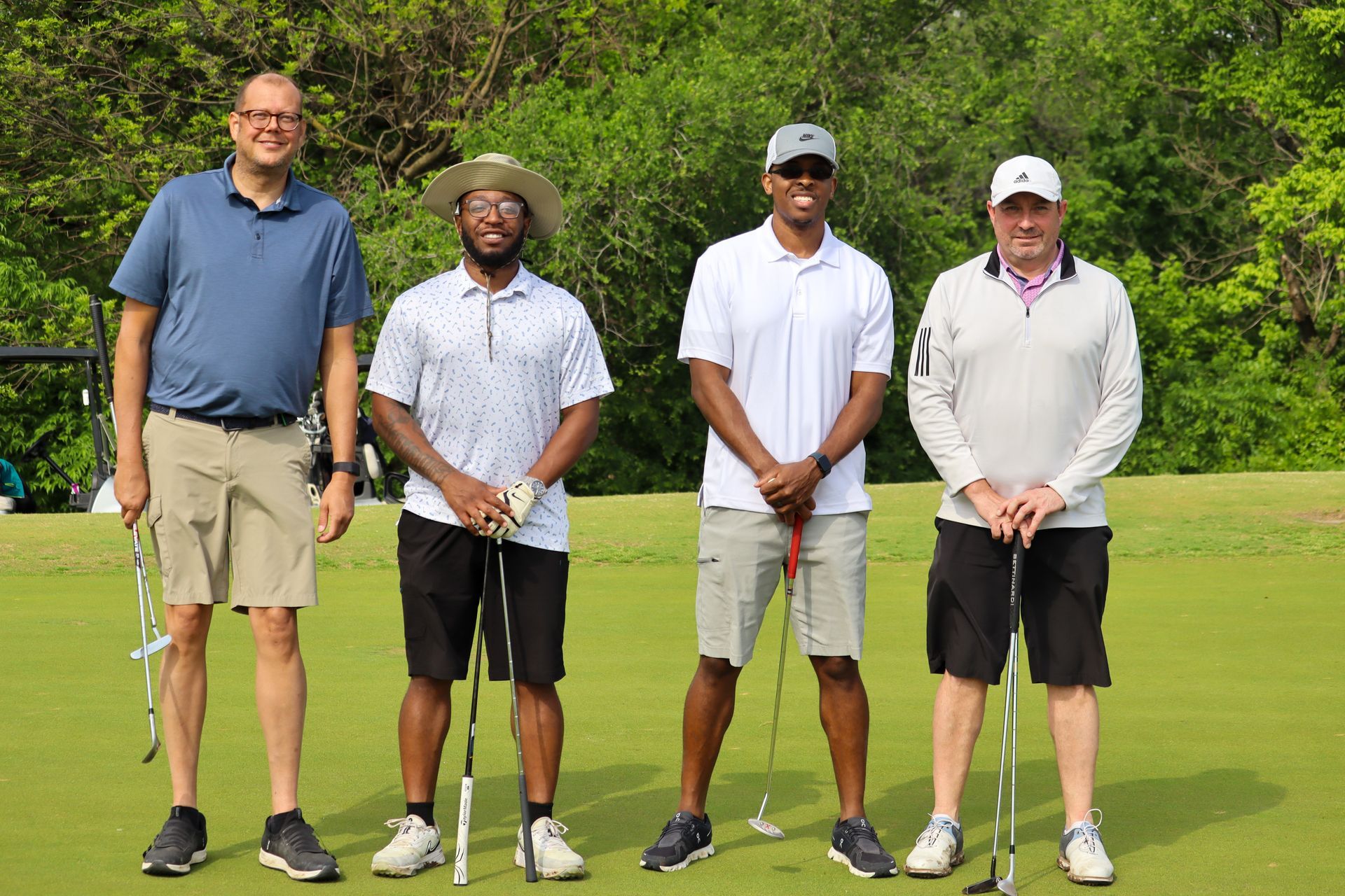 Four men standing on a golf course, holding clubs, smiling; green grass and trees in background.