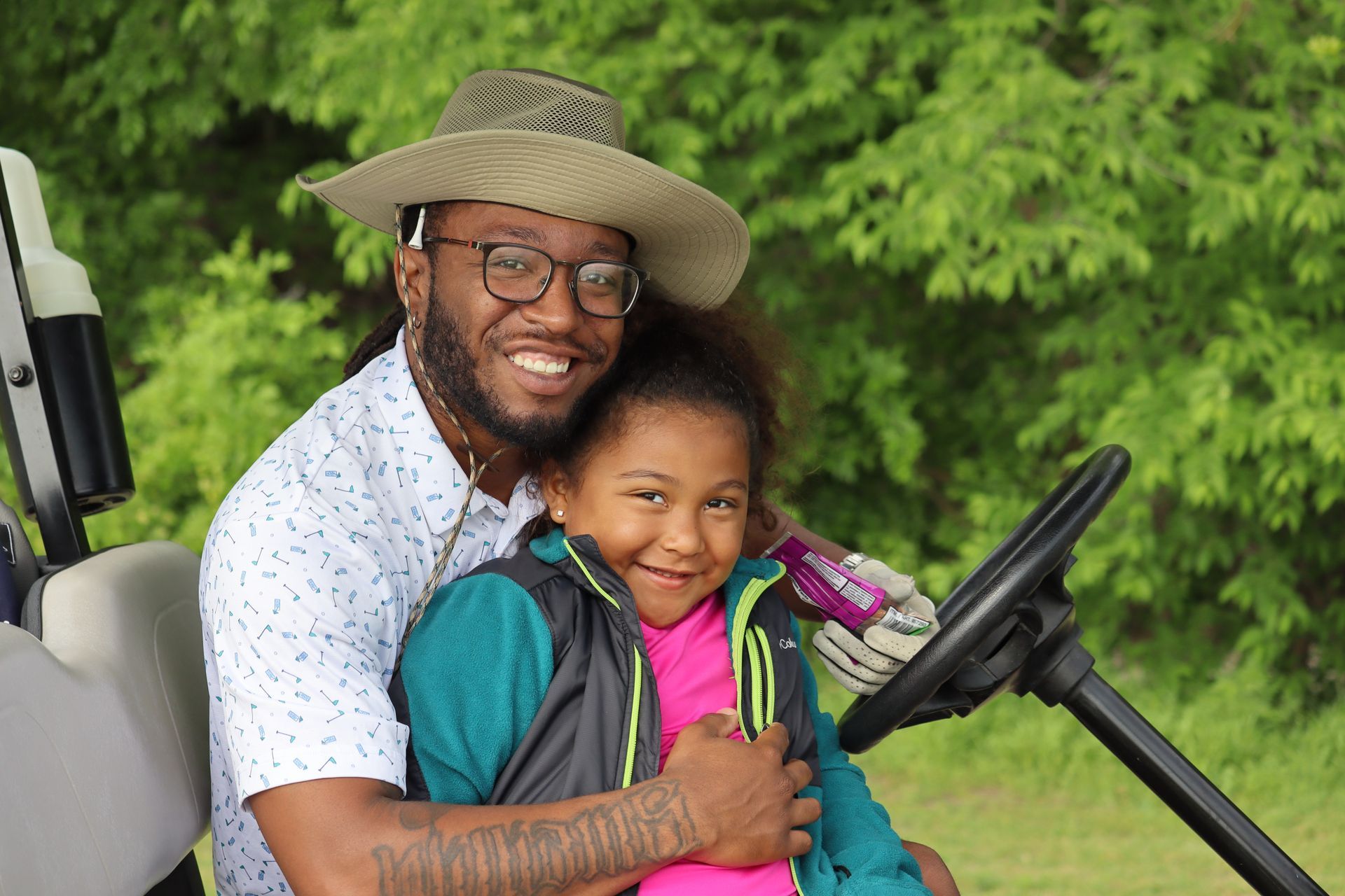 Man and young girl smiling on a golf cart, outdoors. Man wears hat and glasses, girl has a jacket.