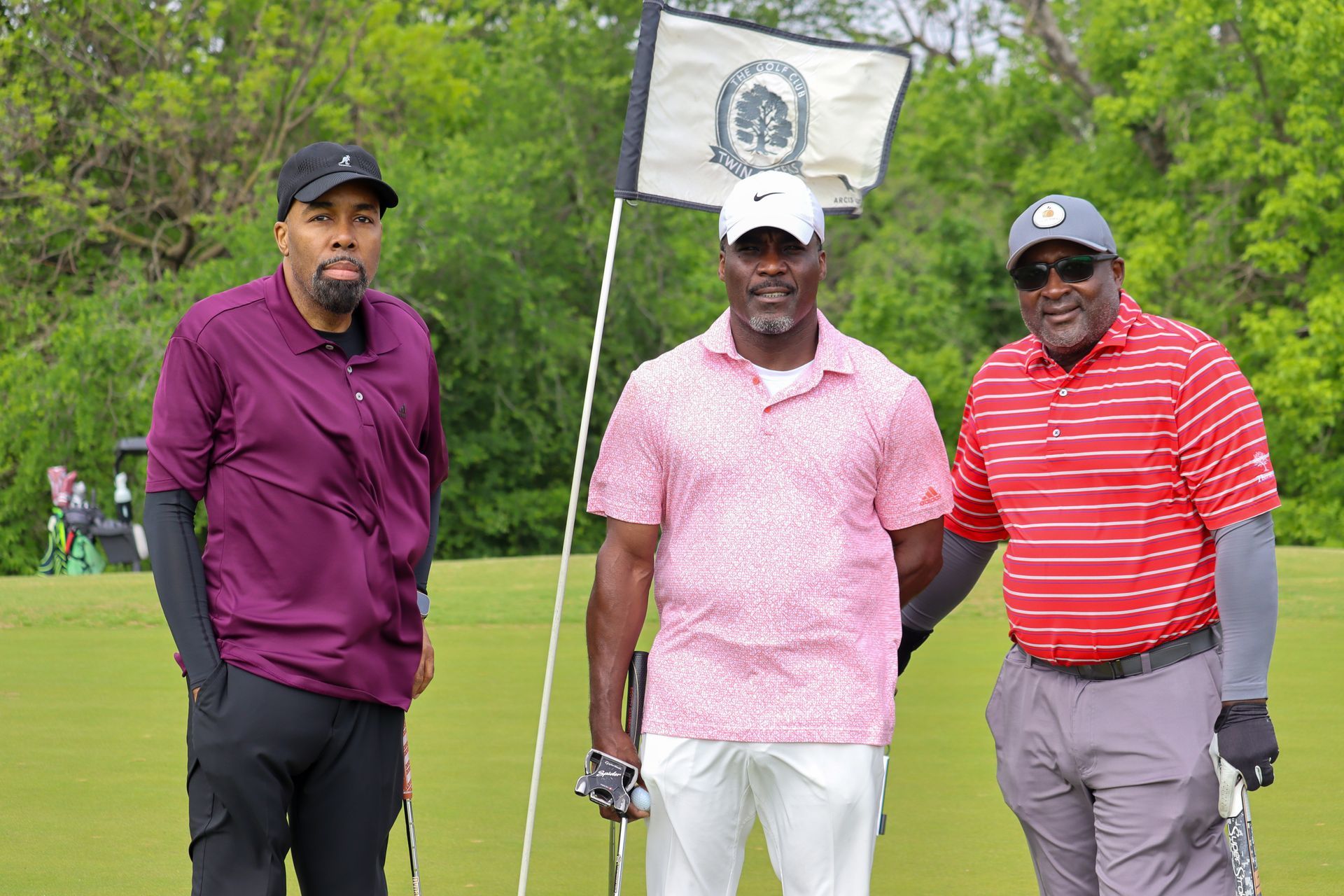 Three men posing on a golf course. Two are holding clubs, all smiling, in colorful attire.