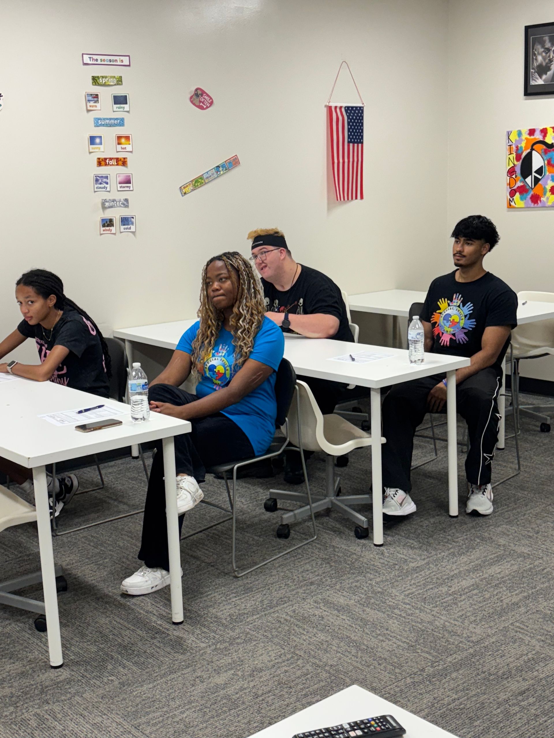 Classroom with four students. One woman with curly blonde hair and three men. One has a colorful design t-shirt.