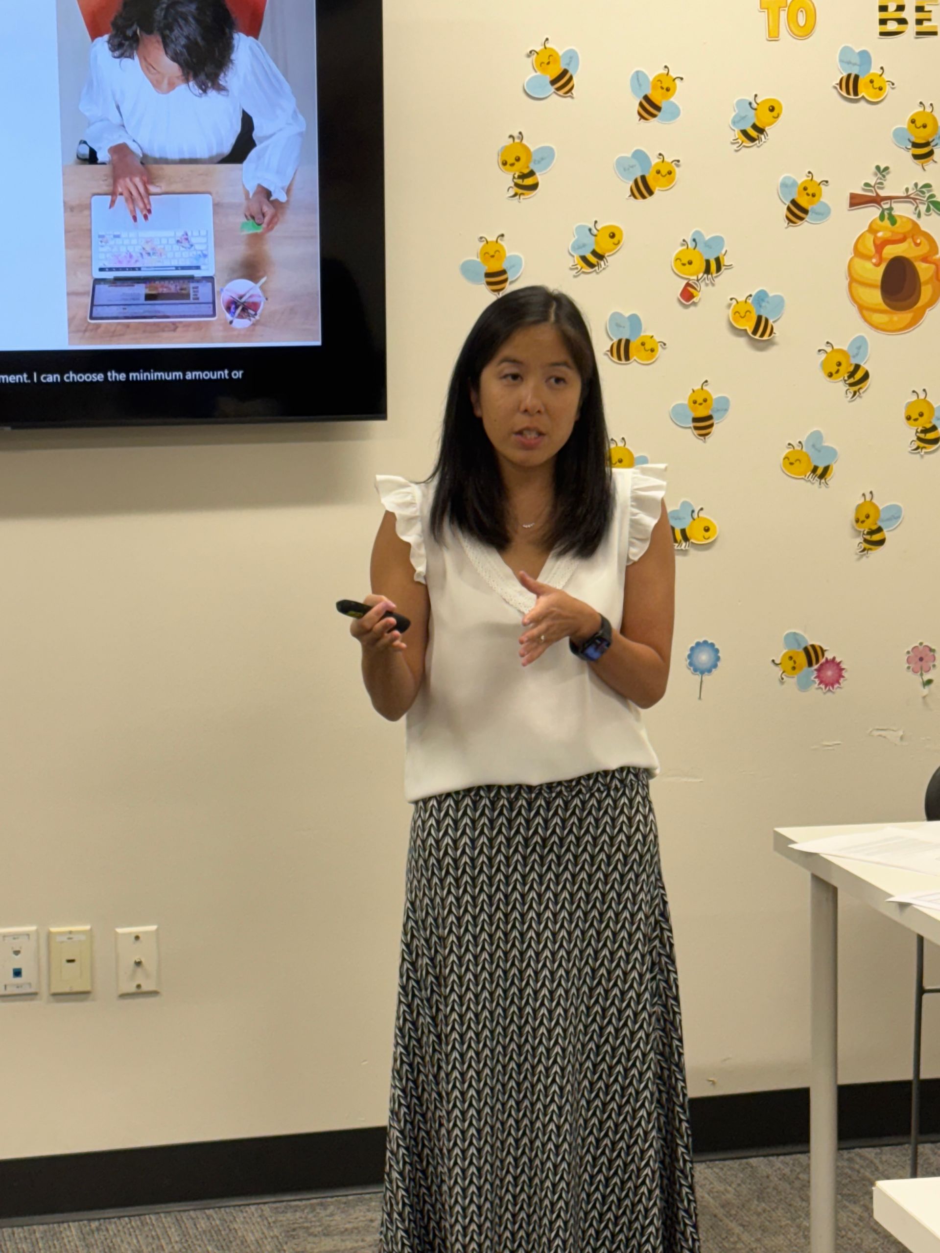 Woman presenting, holding a pointer, in front of a screen and bee-themed wall decorations.