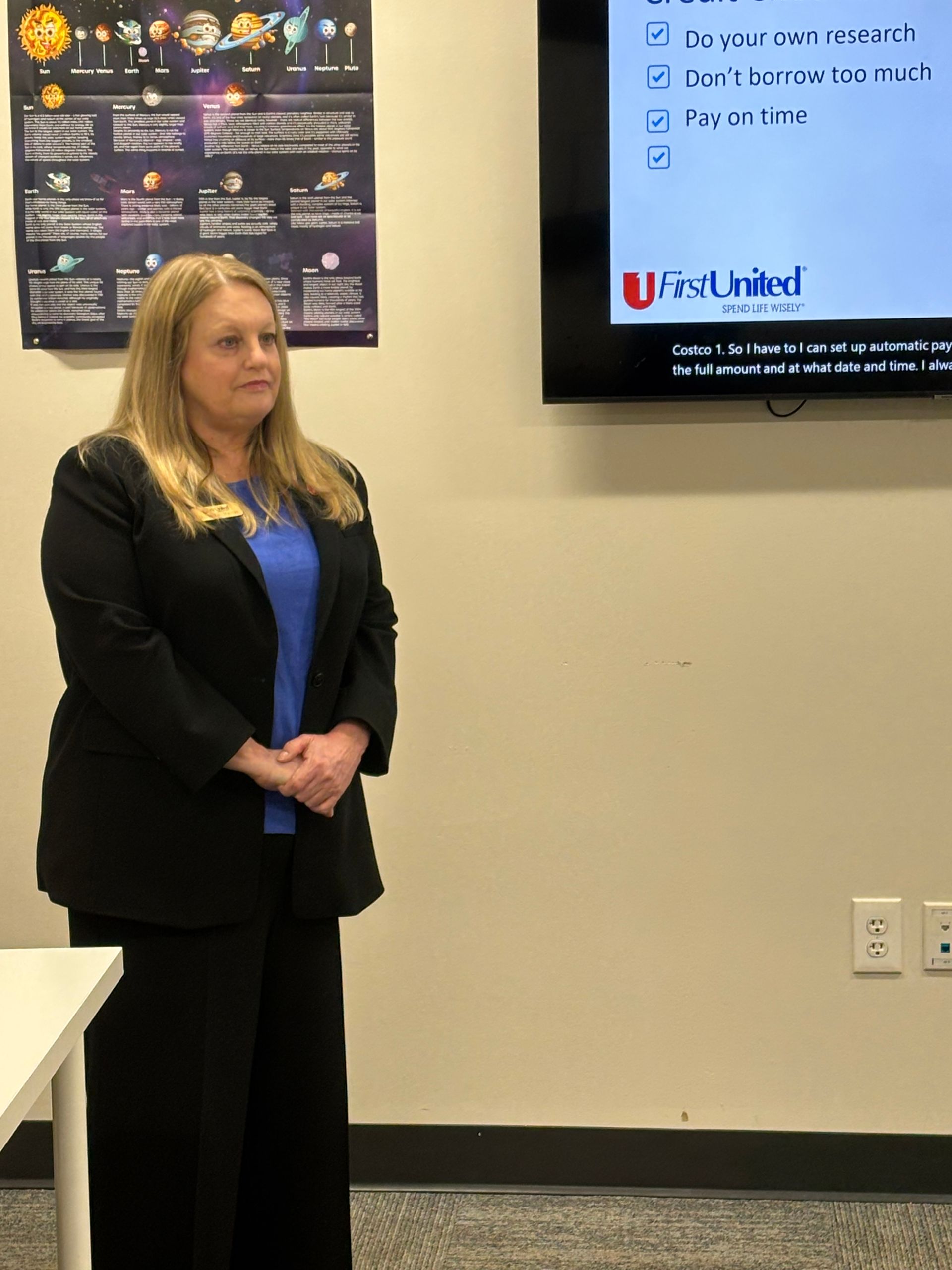 Woman in a black blazer and blue shirt stands near a screen with text, in a classroom setting.