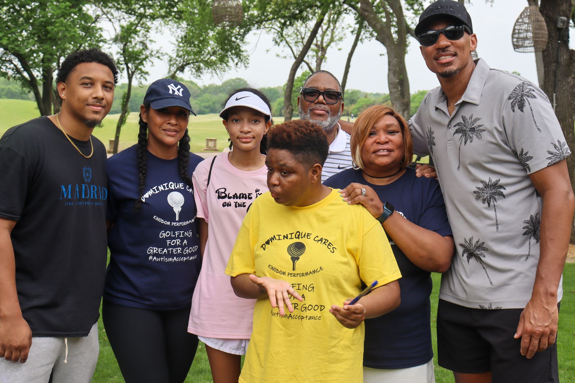 Group of people smiling outdoors, some wearing matching yellow or blue t-shirts; green grass, trees in background.