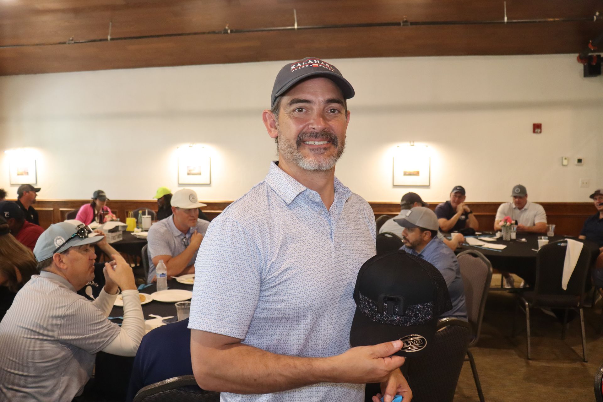 Man in a patterned shirt and cap, holding a black cap, smiles in a room with tables and people.