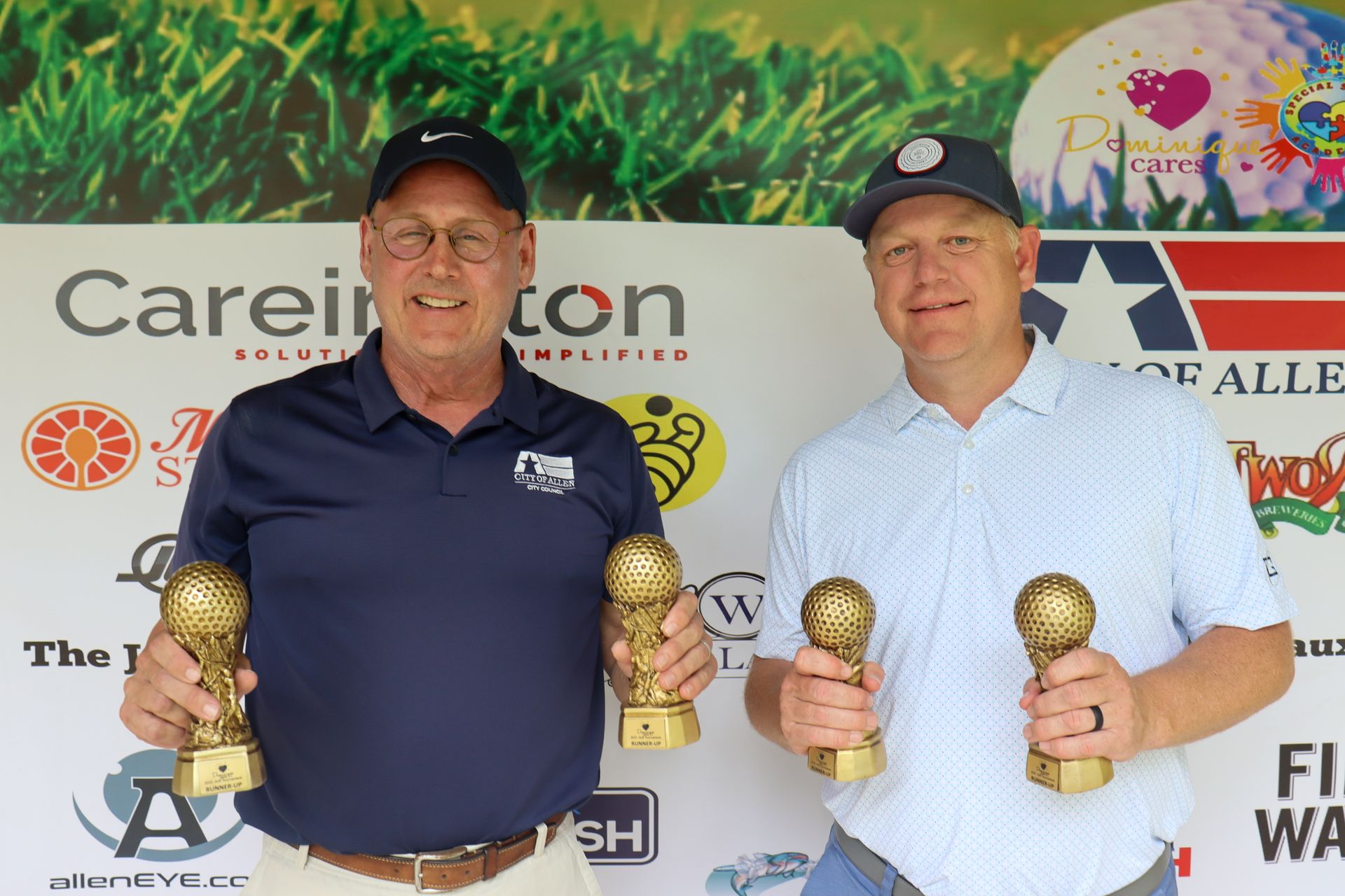 Two men holding trophies at a golf event. They are smiling and posed in front of a sponsor backdrop.