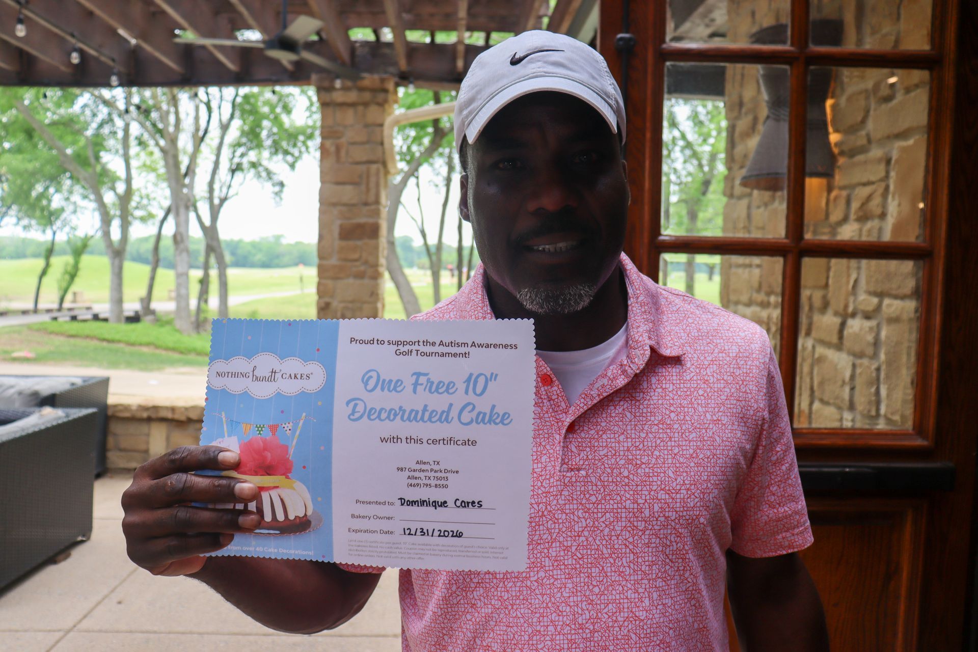 Man holding a certificate for a decorated cake, smiling outdoors. He wears a pink shirt and a white cap.