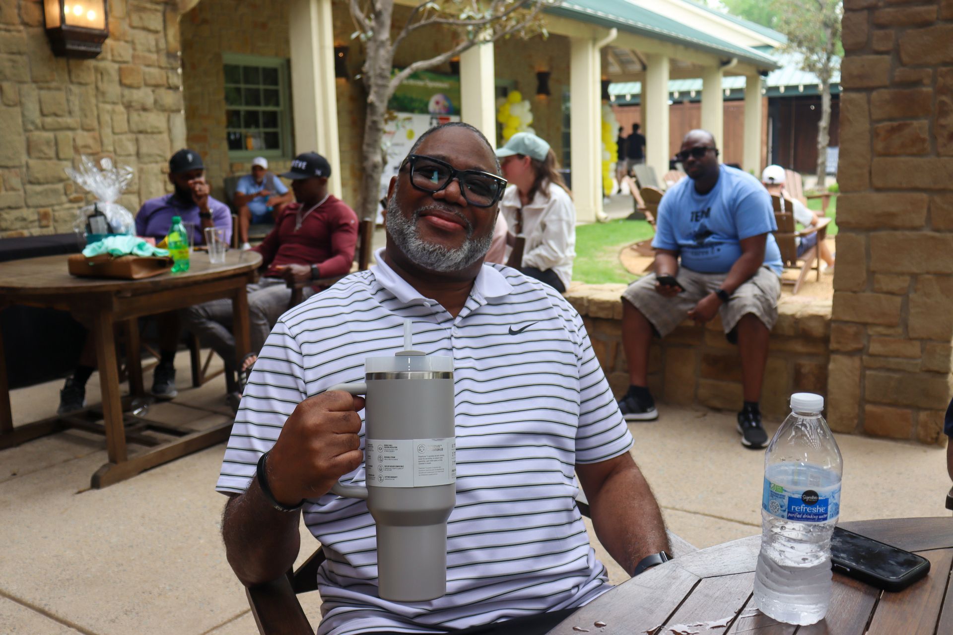 Man smiling, holding a large gray tumbler, seated outside at a table with others; water bottle nearby.