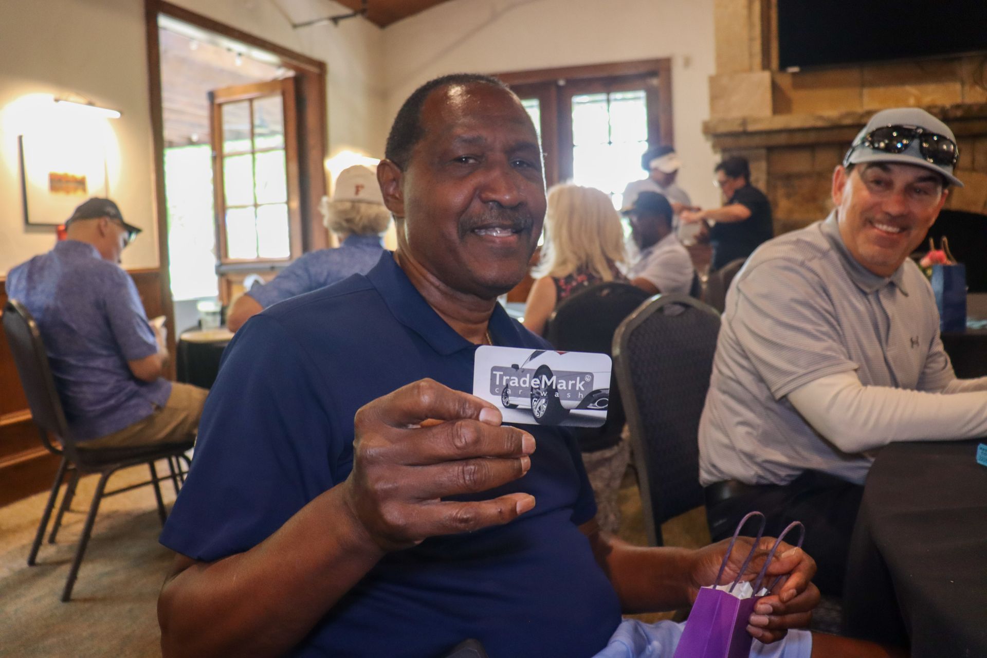 Man holding a small card, smiling. Indoors, people in background, seated around tables.