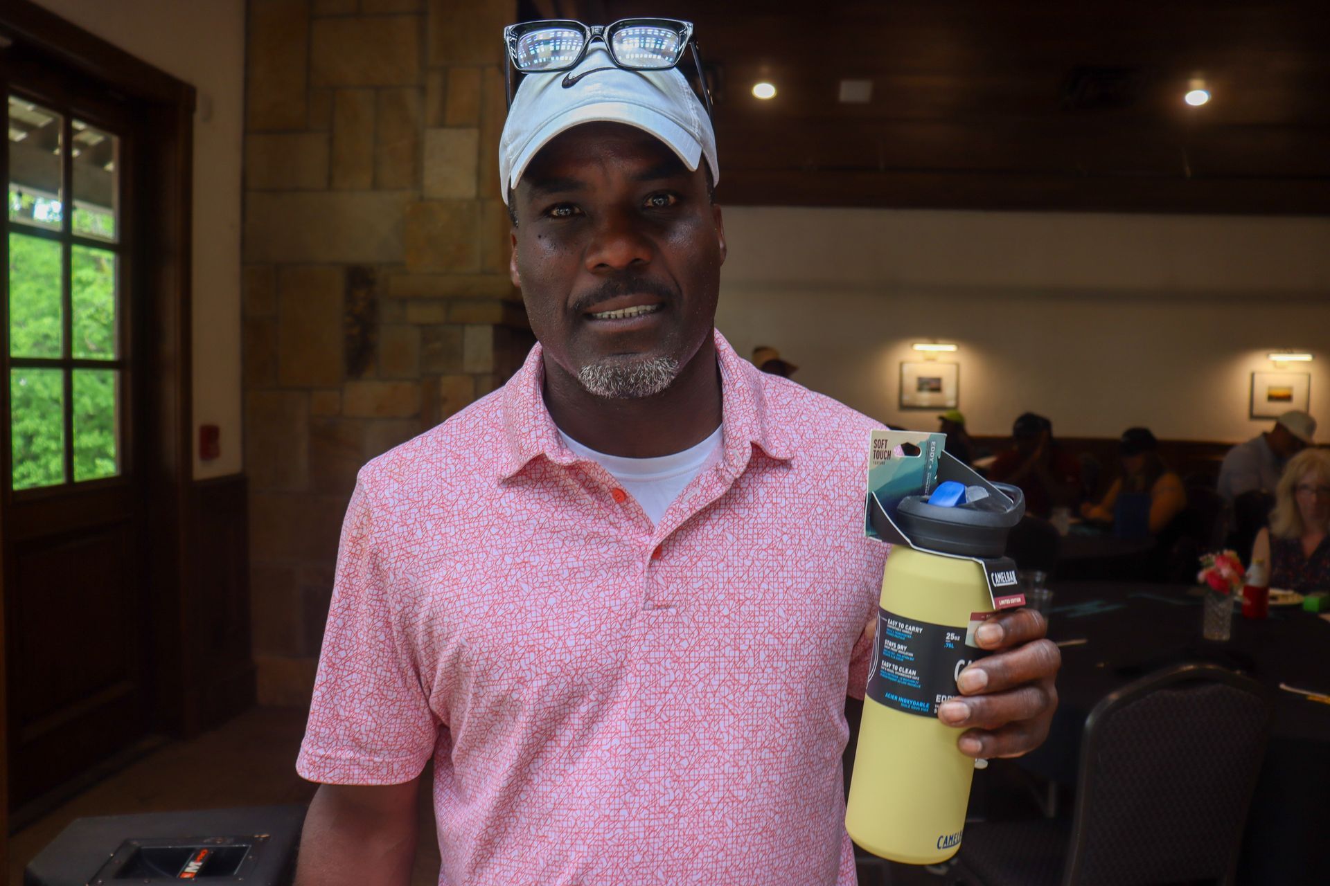 Man in pink shirt, holding a yellow water bottle indoors. He wears a cap and glasses.