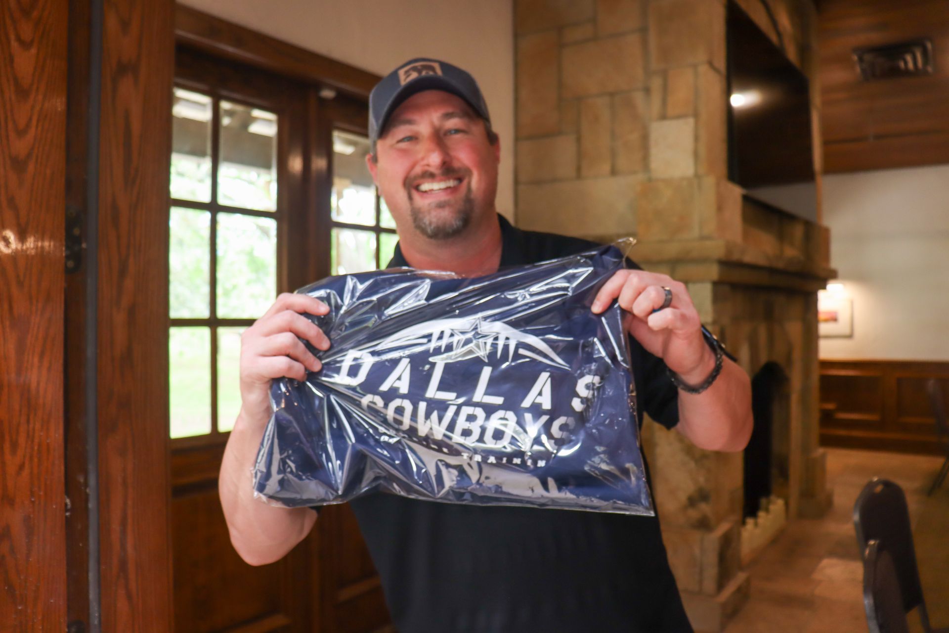 Man smiles, holding a sealed Dallas Cowboys shirt in a room with a fireplace.