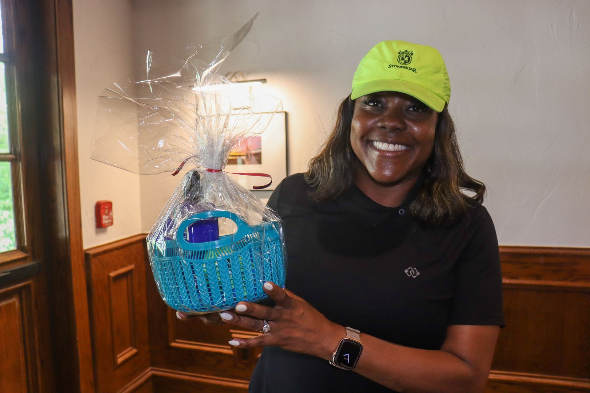 Woman in a yellow cap smiles, holding a blue gift basket indoors near a window.