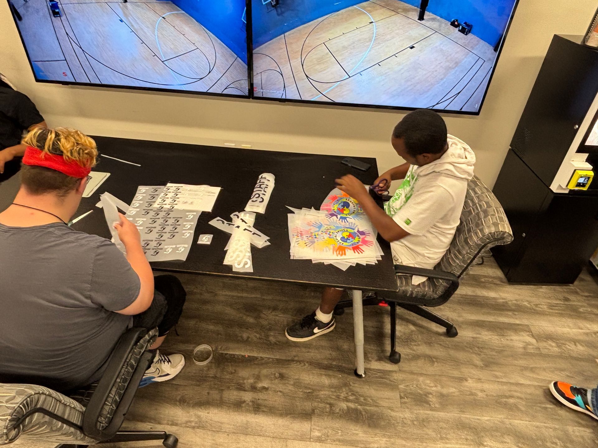Two people seated at a table assembling paper items. Two screens display a basketball court.