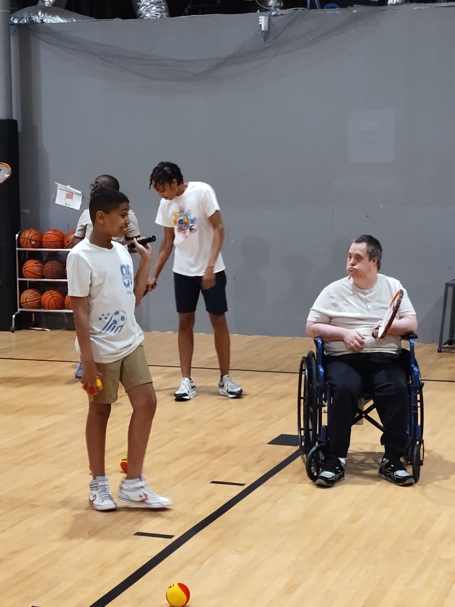 Basketball court, three people: boy with ball, teen, man in wheelchair with racket.