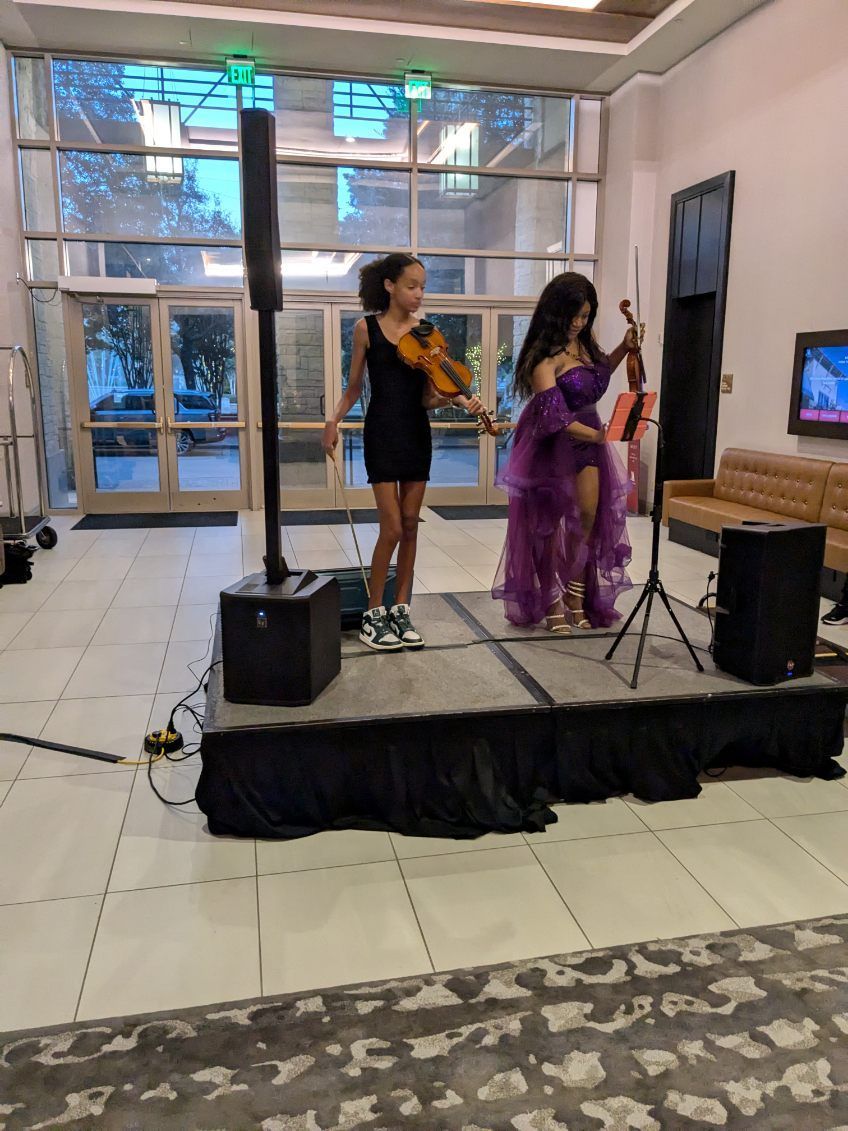 Two violinists performing on a stage in front of a glass wall, one in black and one in purple.