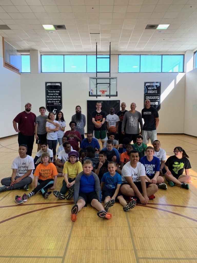Group of kids and adults in a basketball court. Some are sitting, some standing, with basketball and hoop.