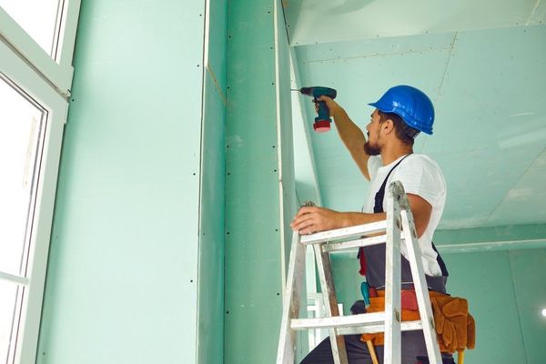 Worker in blue hard hat drilling a wall beside a ladder in a light green room