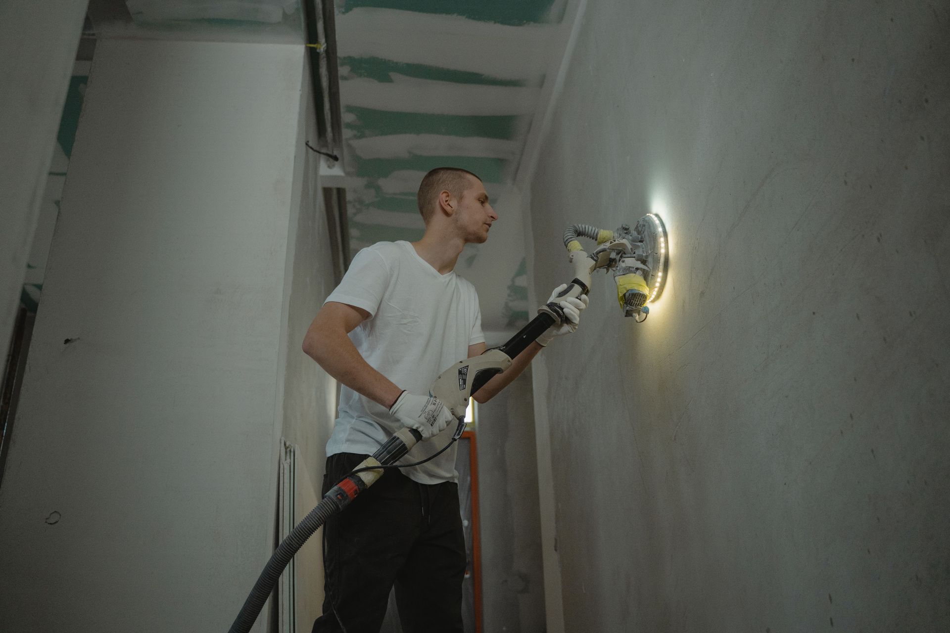 Worker sanding a wall with a power tool in a narrow hallway under construction