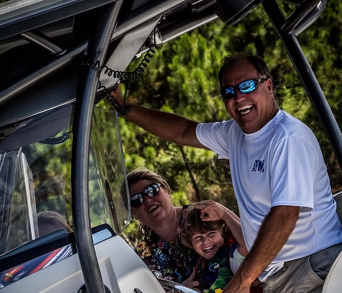 Family on a boat, smiling and laughing. Sunlight, blue sunglasses, and coastal setting.