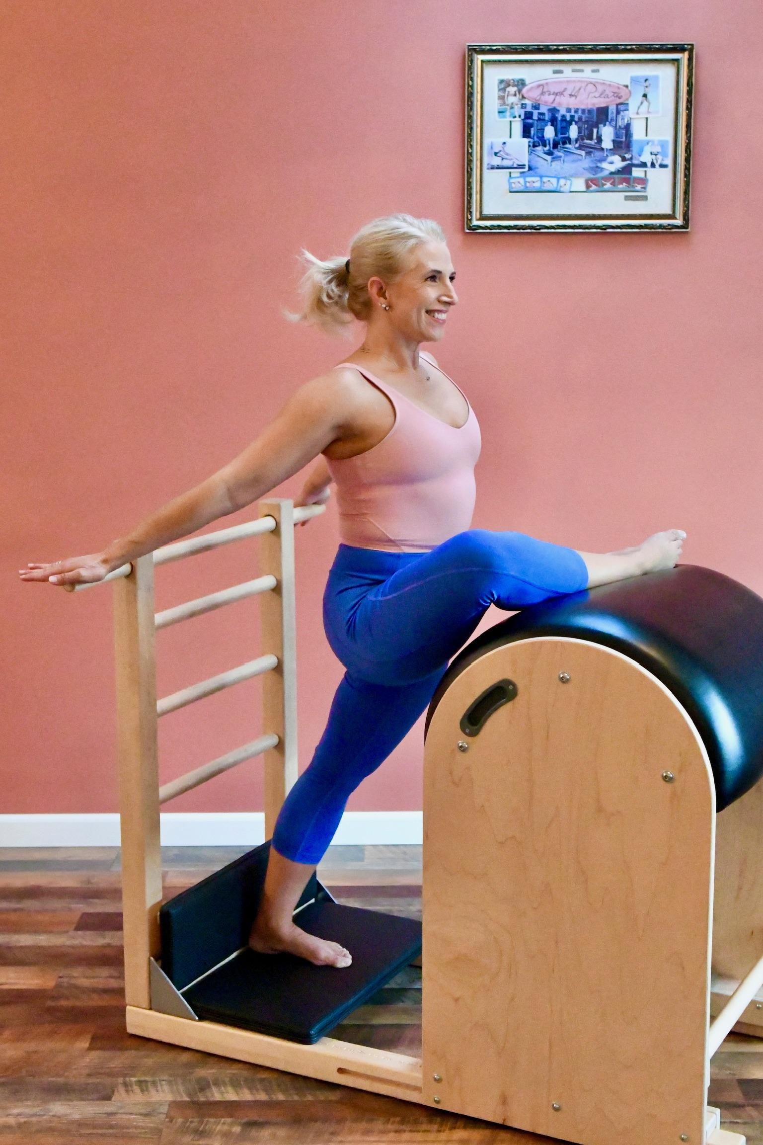 Woman exercising on a Pilates reformer, balancing one leg on a curved barrel in a studio.