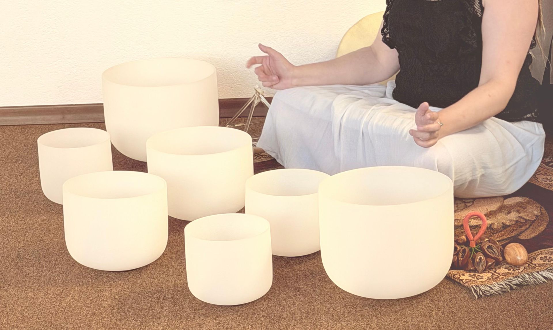 Person meditating among white singing bowls on a woven mat indoors